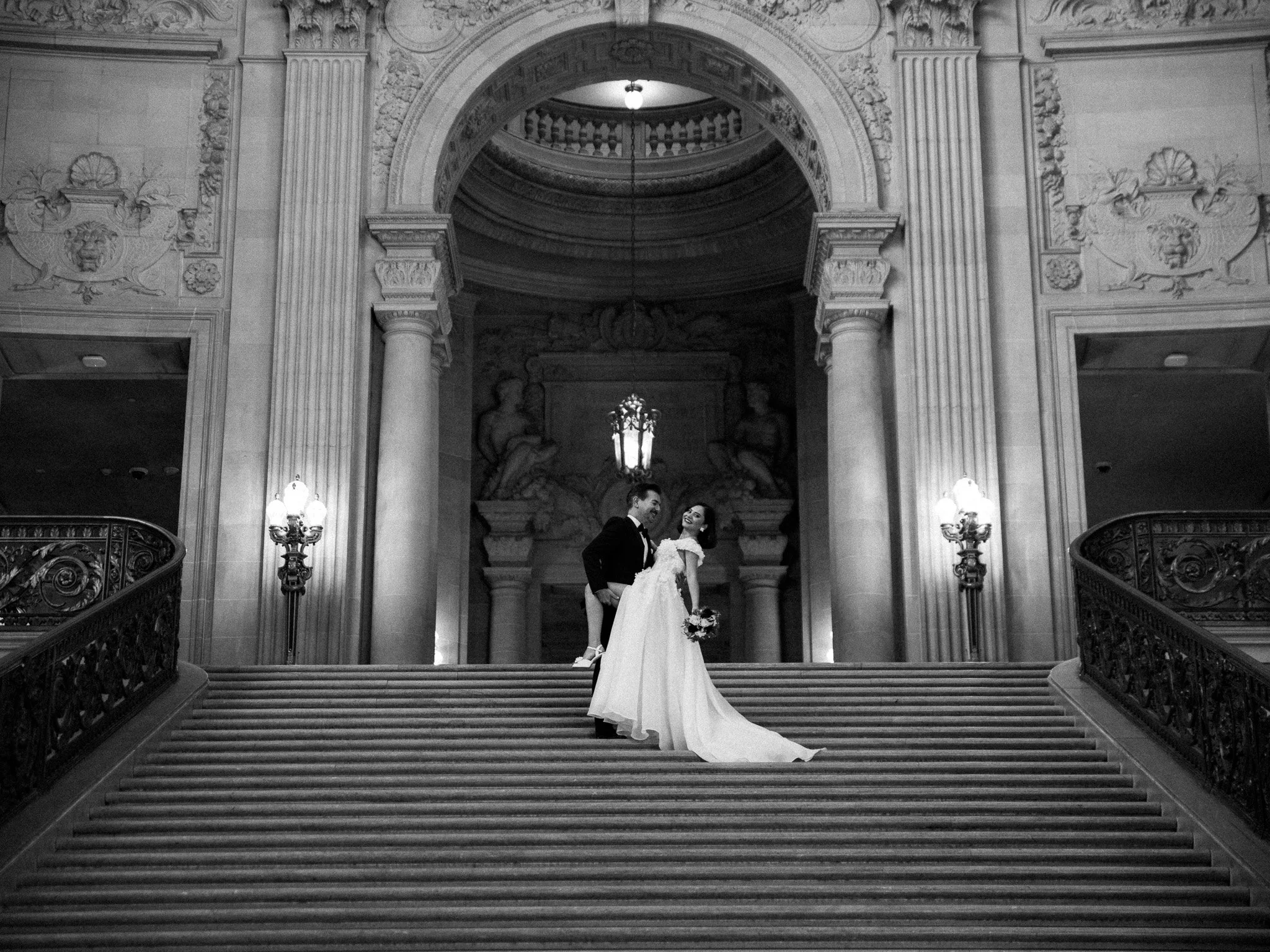 Black and white photo of a bride and groom standing on a grand staircase inside a historic building with ornate architecture, columns, sculptures, and decorative lighting.