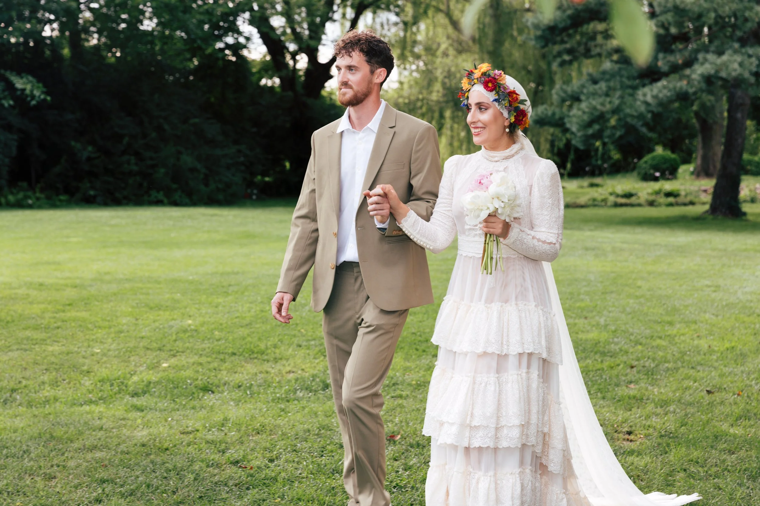 A bride and groom walking outdoors on a grassy area surrounded by trees, with the bride holding a bouquet of flowers and wearing a floral crown, while the groom wears a tan suit.
