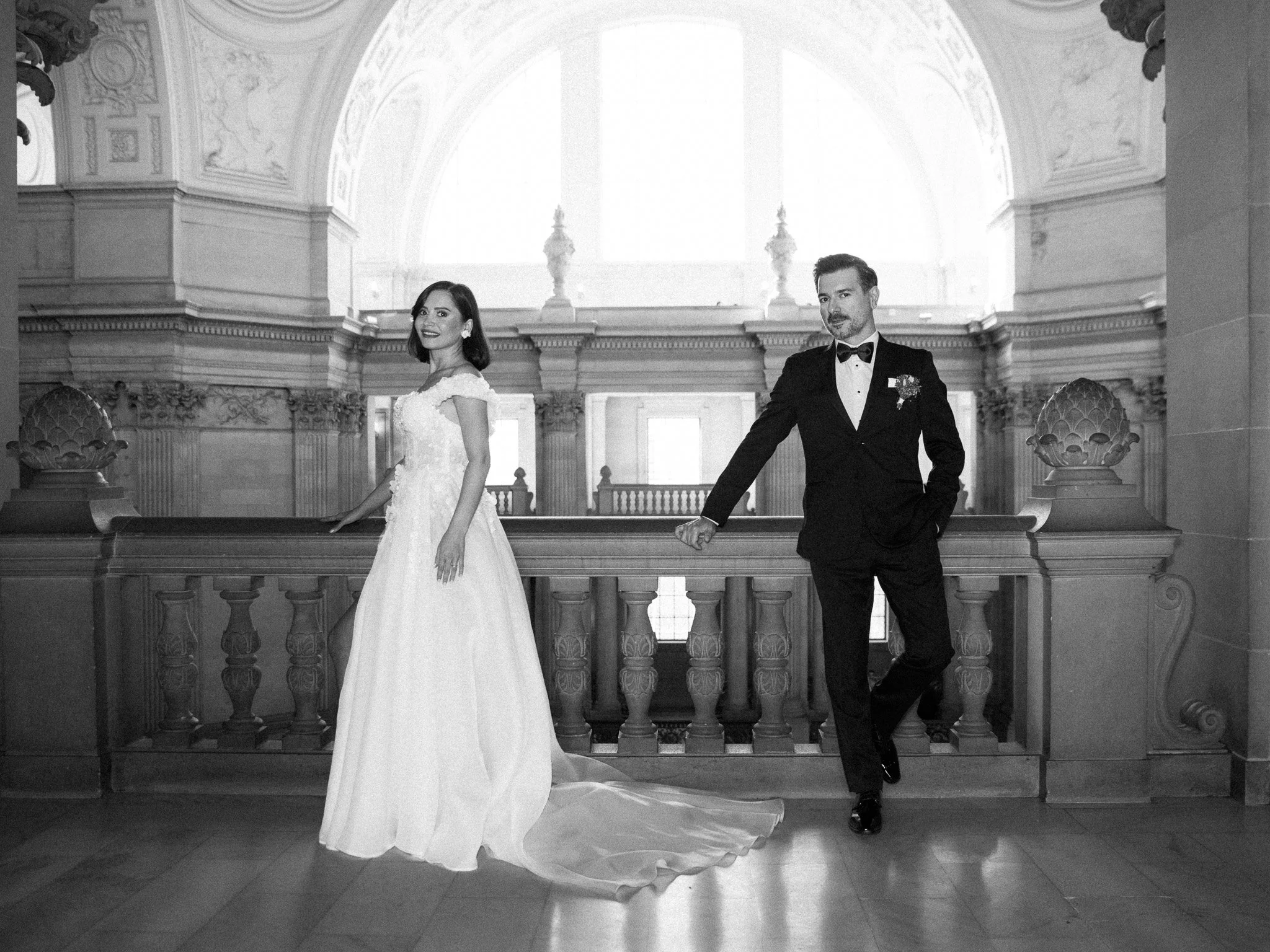 A bride in a wedding gown stands next to a groom in a tuxedo in a grand, ornate hall with high ceilings and decorative architecture.