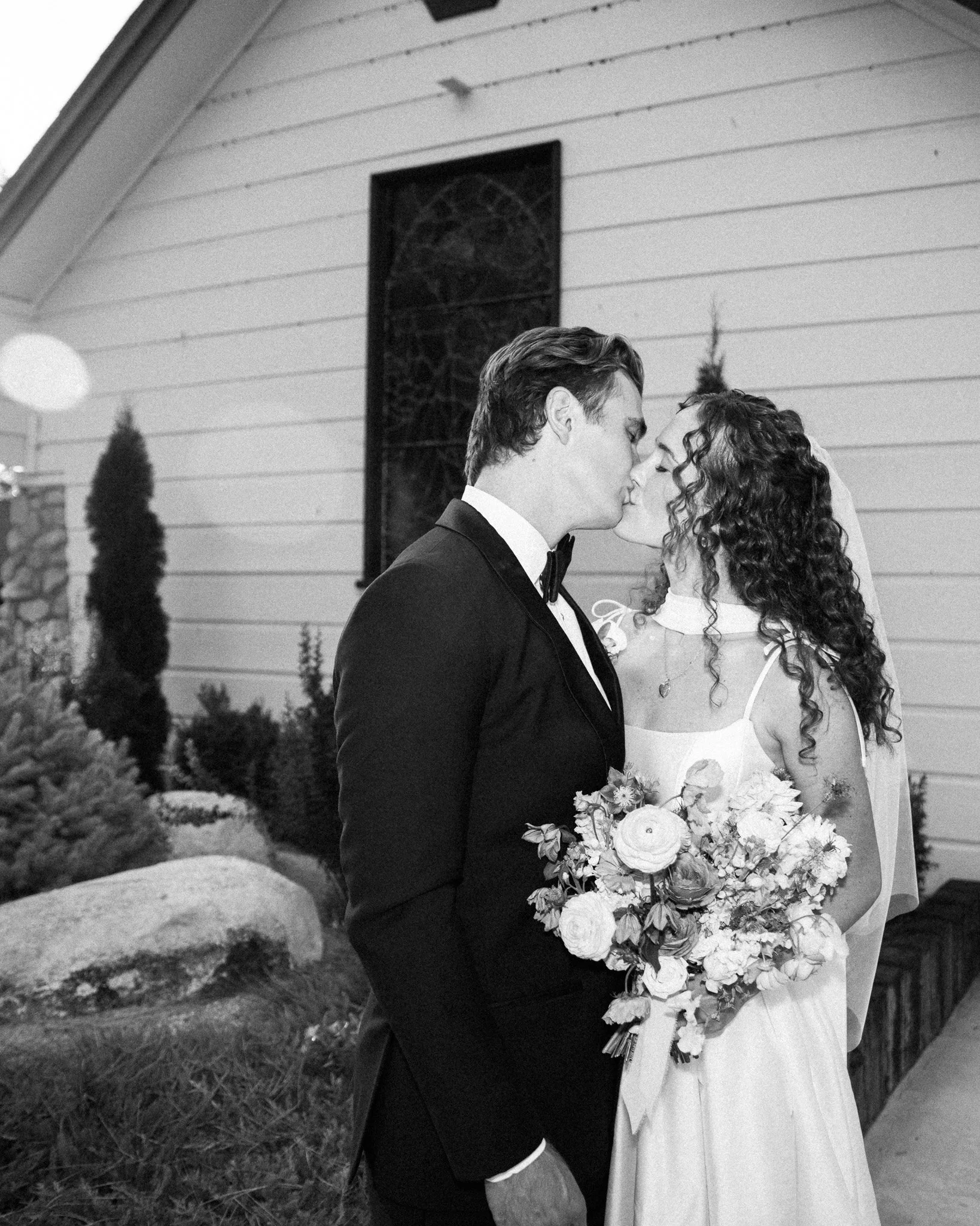 A black-and-white photo of a bride and groom sharing a kiss outside a building with a garden, the bride holding a bouquet of flowers, the groom wearing a tuxedo.