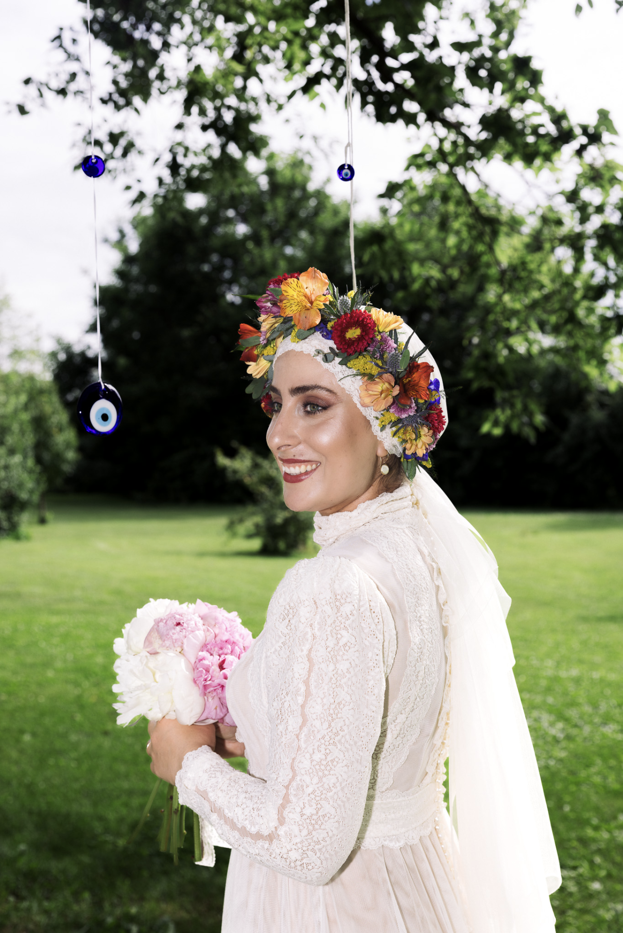 A bride in a lace wedding dress holding a bouquet of pink and white flowers, wearing a floral crown and smiling outdoors with greenery in the background.