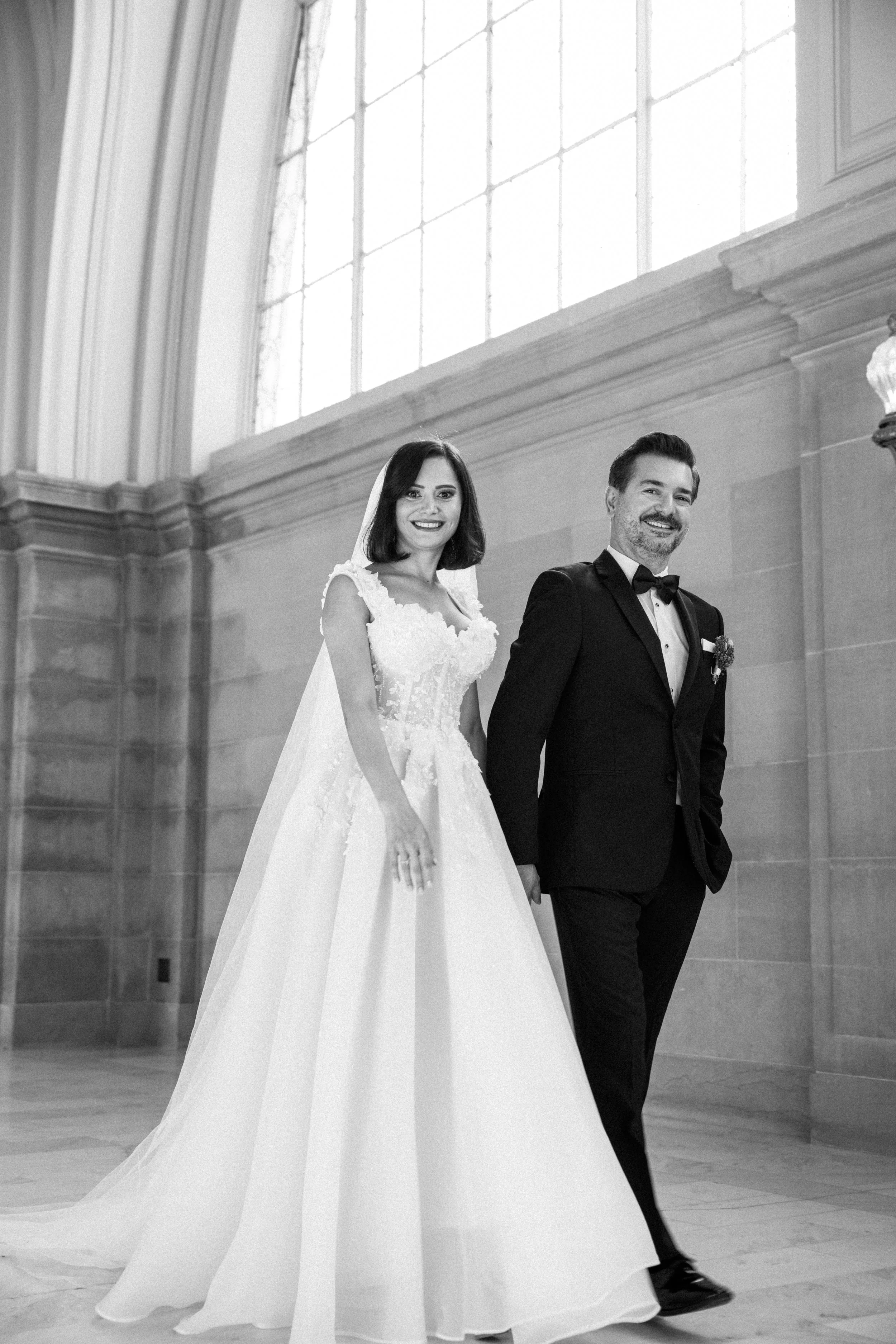 A bride and groom holding hands and smiling indoors, with large windows and classical architecture behind them.
