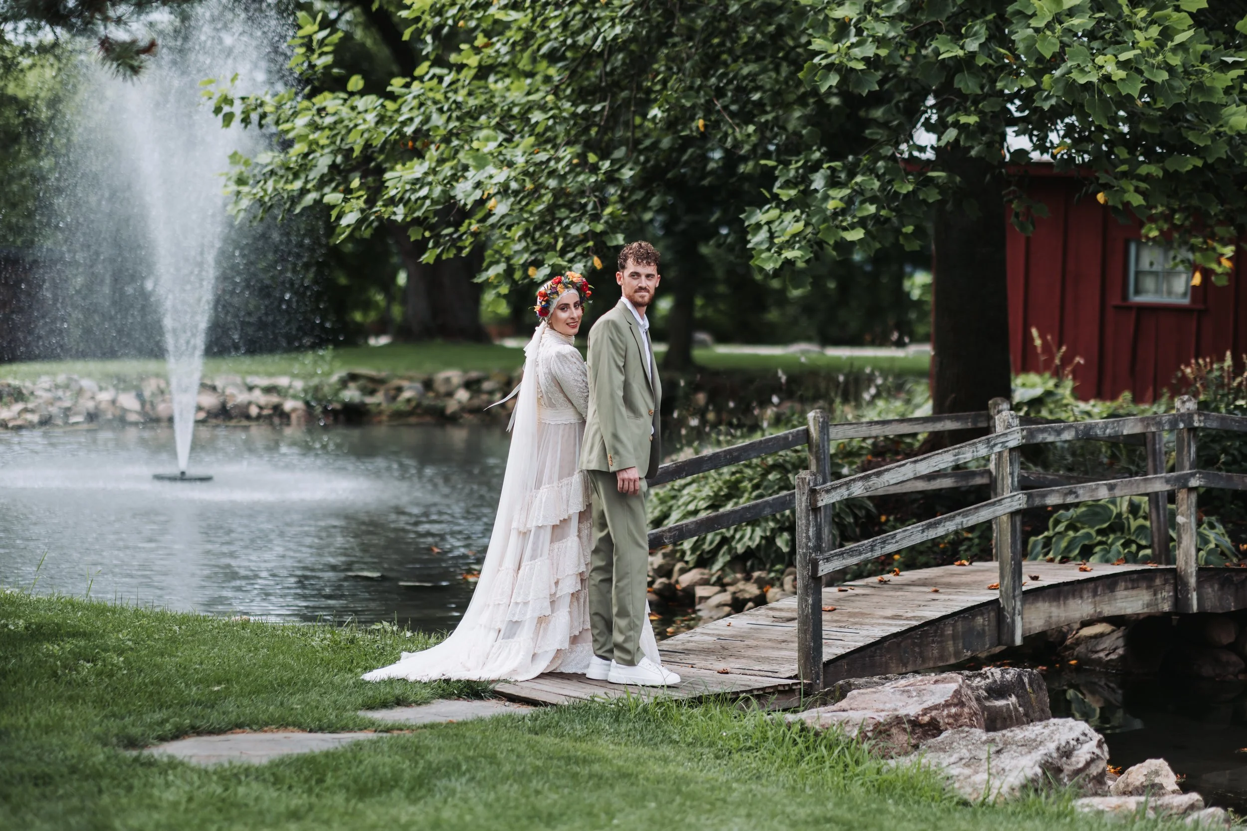 A bride and groom standing on a wooden bridge by a pond in a lush garden. The bride is wearing a long white wedding dress with lace details and a floral headpiece, and the groom is dressed in a light green suit with a white shirt and white shoes.