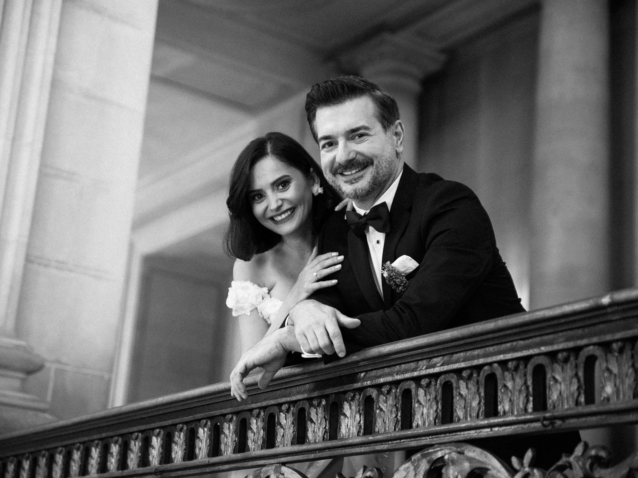Black and white photo of a smiling couple in formal wedding attire, leaning on a decorative wooden railing inside a building with classical architectural elements.