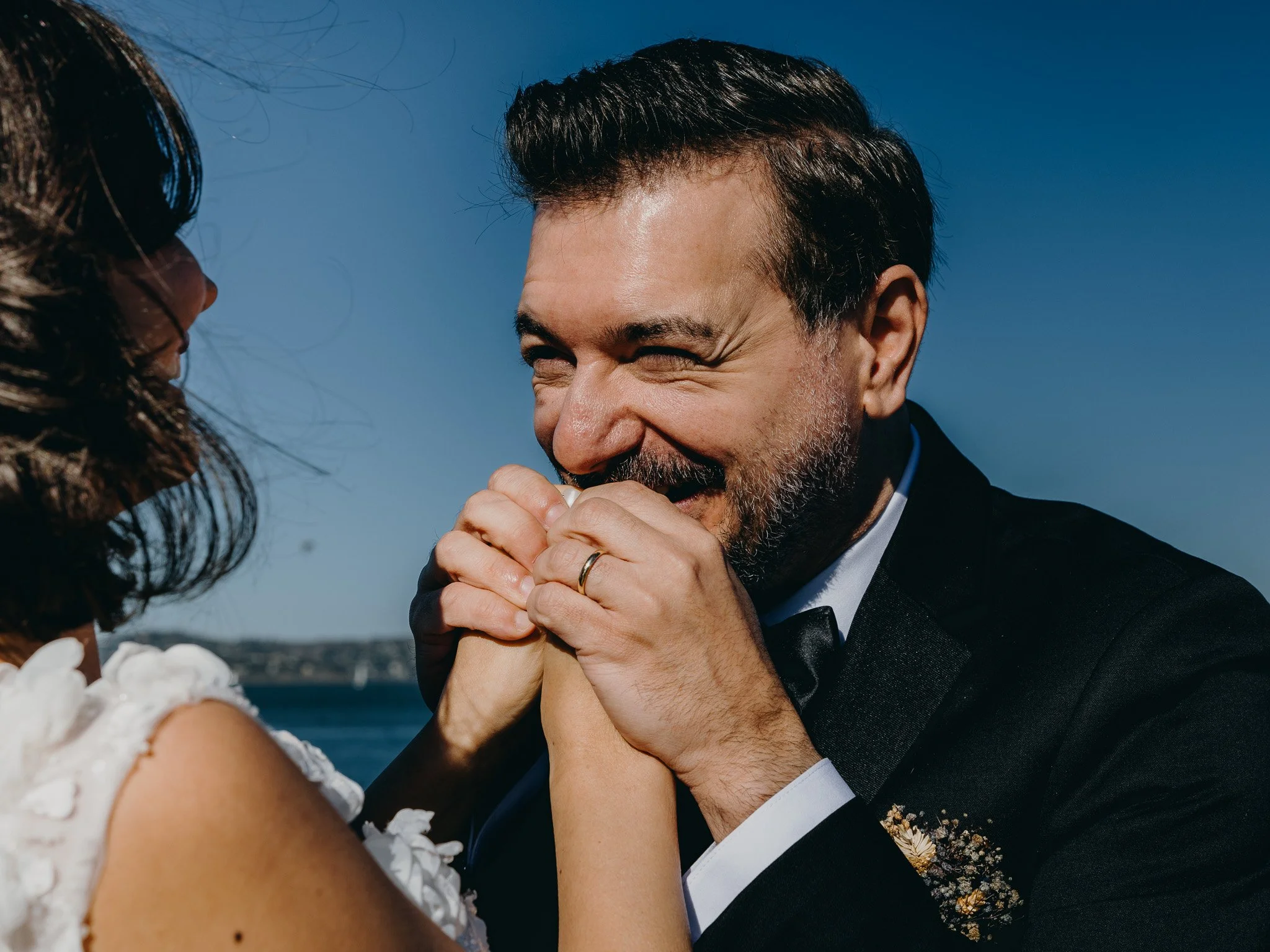 A man smiling and holding a woman's hands during a wedding or romantic moment outdoors with a blue sky background.