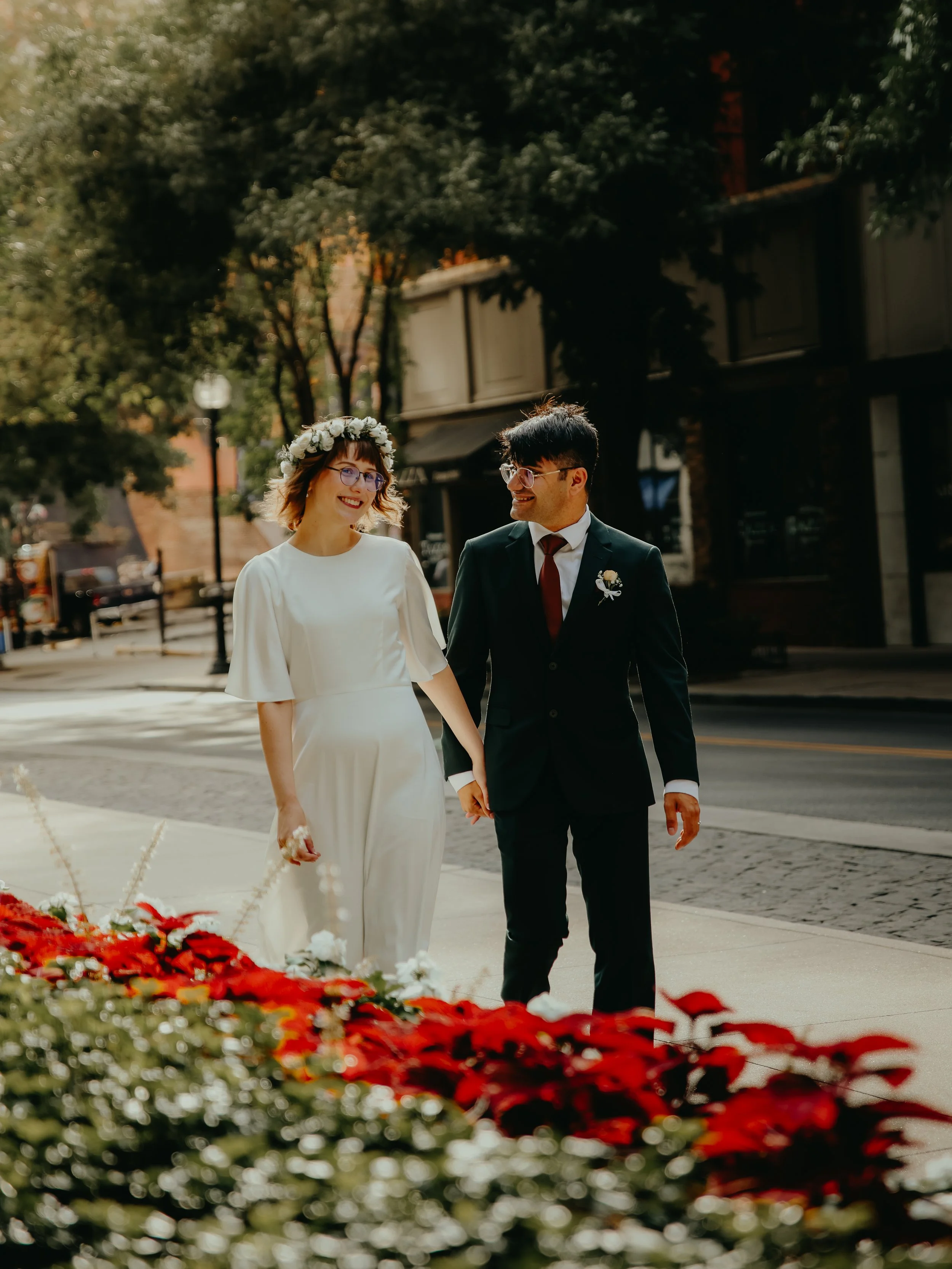 A couple dressed in wedding attire walking hand in hand outdoors on a city sidewalk, surrounded by red and white flowers, with trees and buildings in the background.