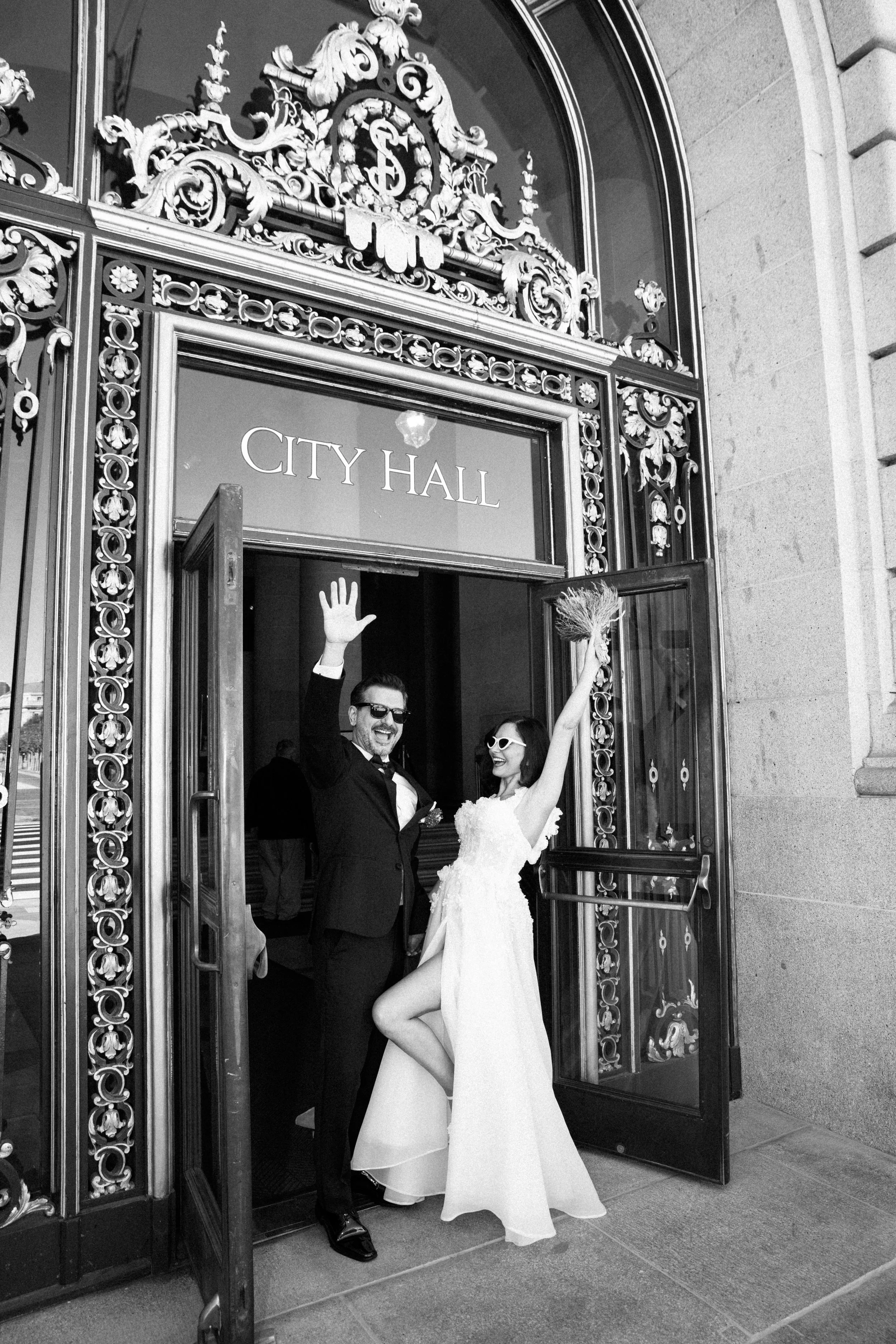 A newlywed couple exiting a city hall building, both waving and smiling, with the woman holding a bouquet in her raised hand.