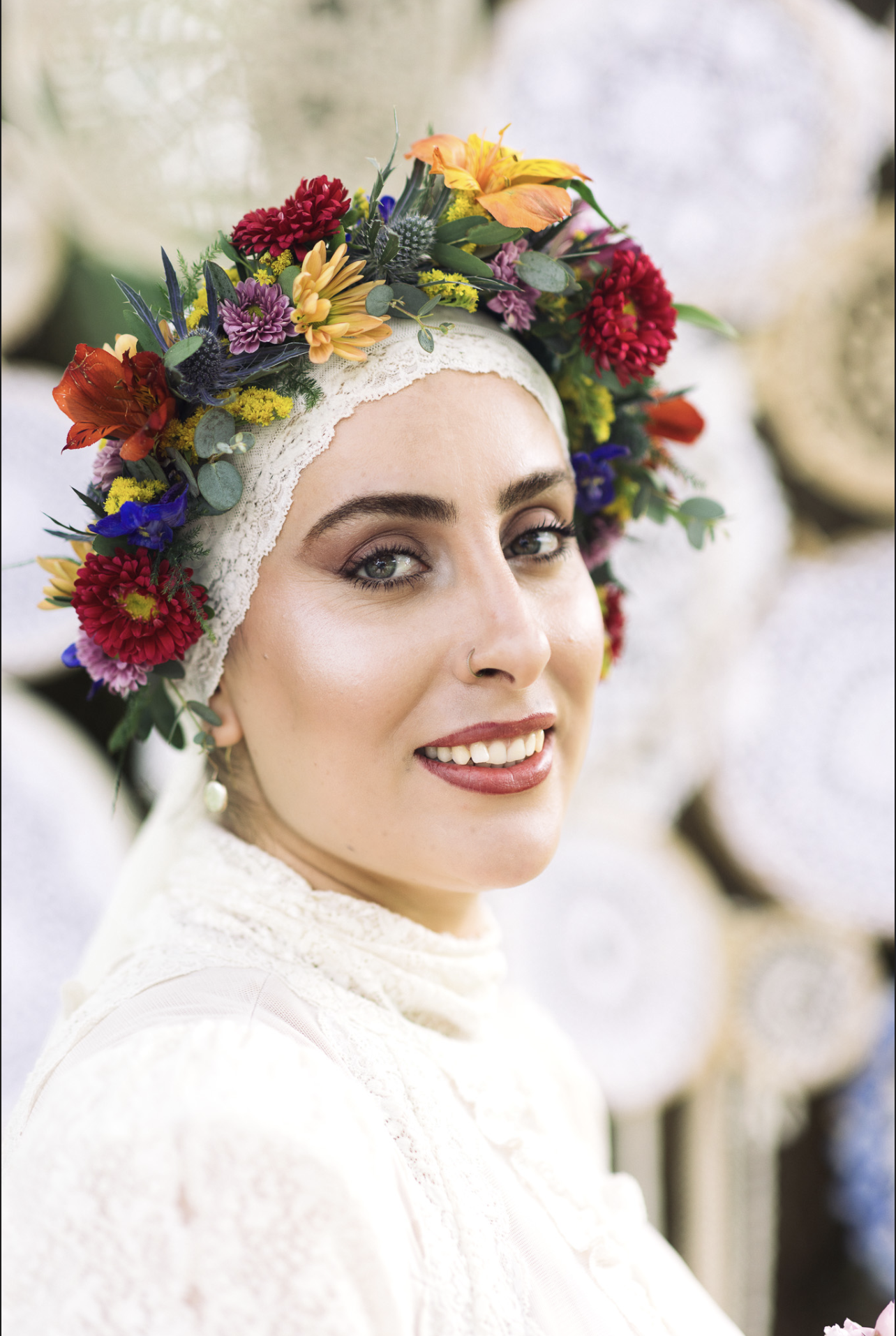 A woman wearing a white lace headwrap decorated with colorful flowers, including red, yellow, purple, and blue, smiling with light makeup and a small nose piercing.