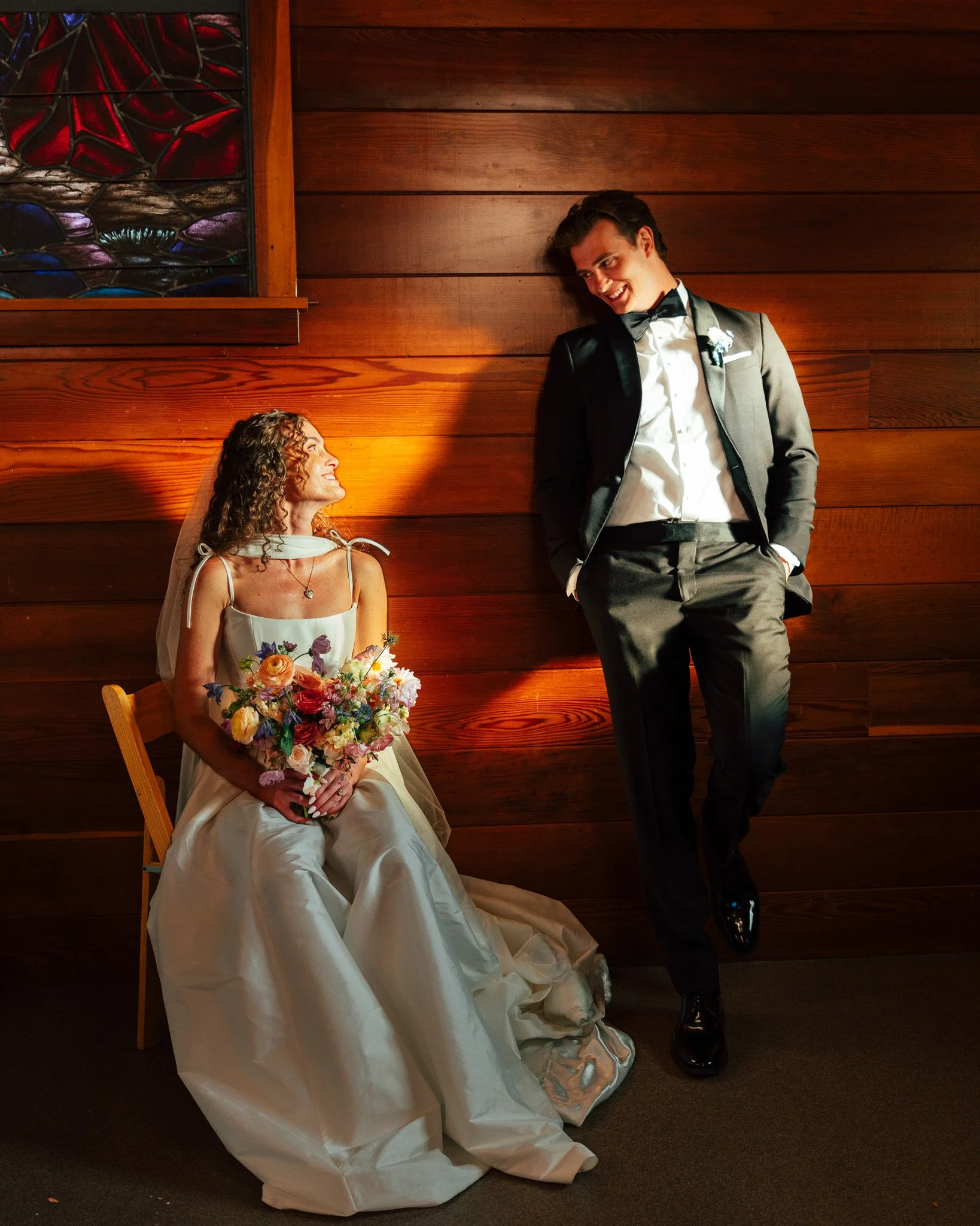 A bride in a wedding dress holding a bouquet of colorful flowers, sitting on a chair, looking up and smiling at a groom in a black tuxedo with a bow tie, who is leaning against a wooden wall and smiling back.