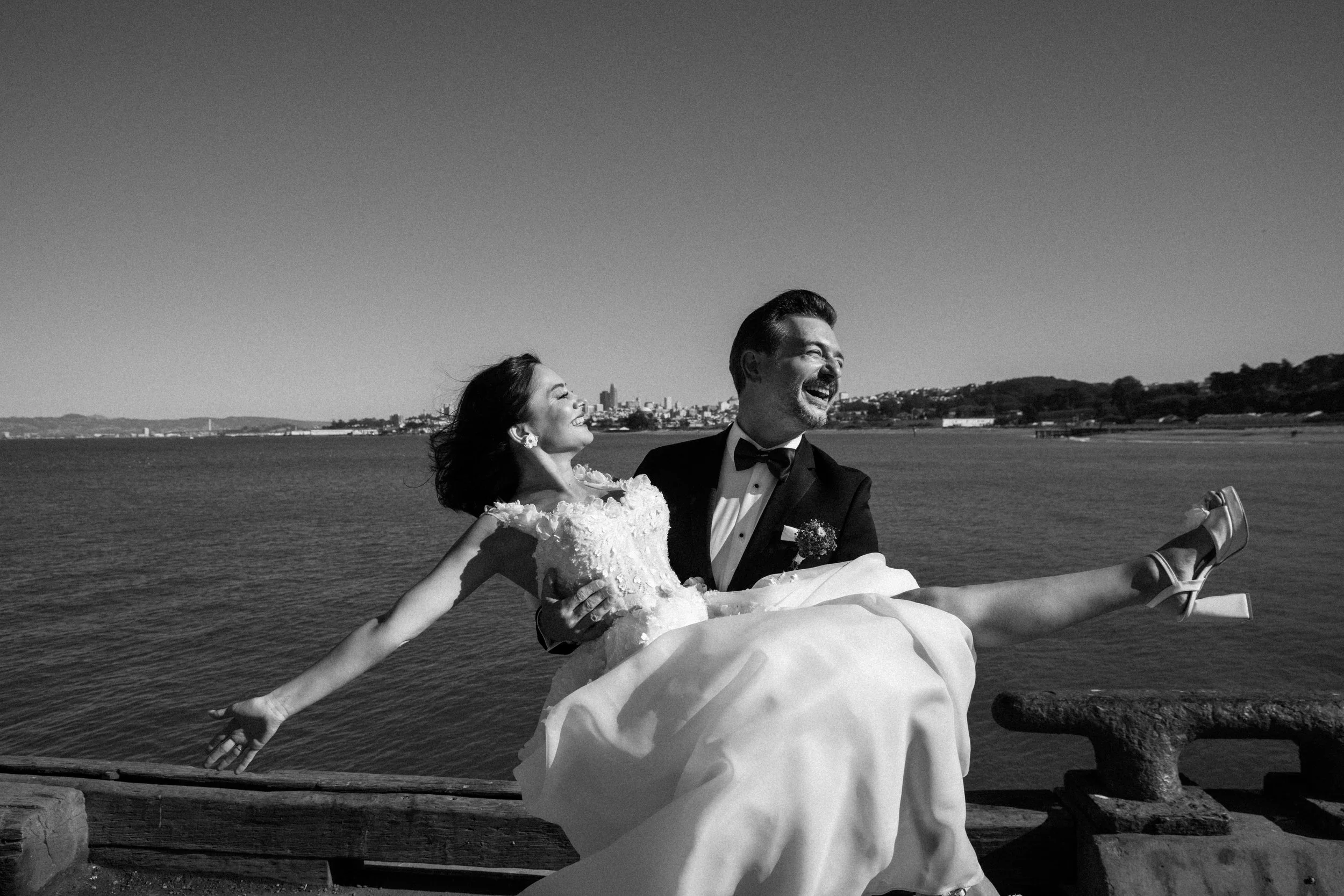 A black-and-white photo of a wedding couple by the water. The groom is holding the bride in his arms, both are smiling and looking at each other. The background includes a body of water and a city skyline in the distance.