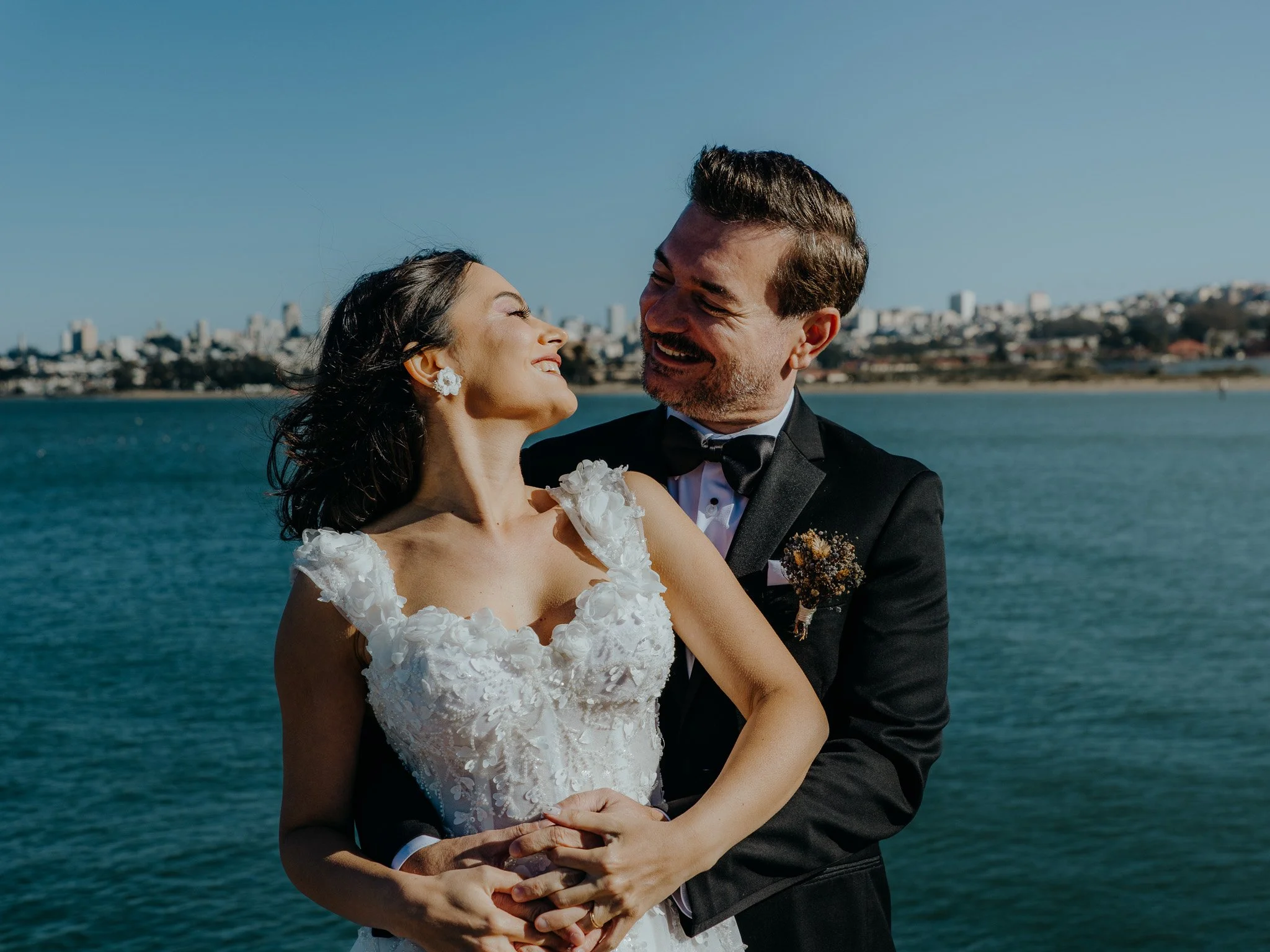 A bride and groom smiling and embracing near a body of water on a sunny day.