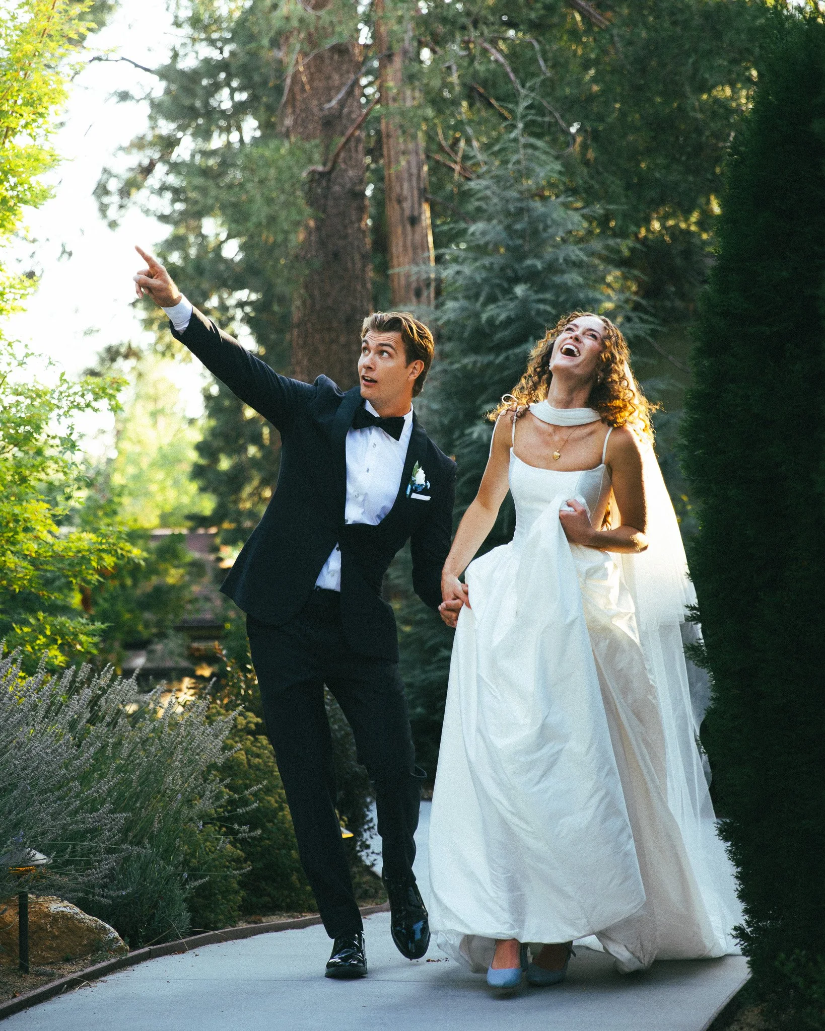 A bride and groom walking outdoors in a lush green garden, holding hands, with the groom pointing upward and both laughing joyfully.