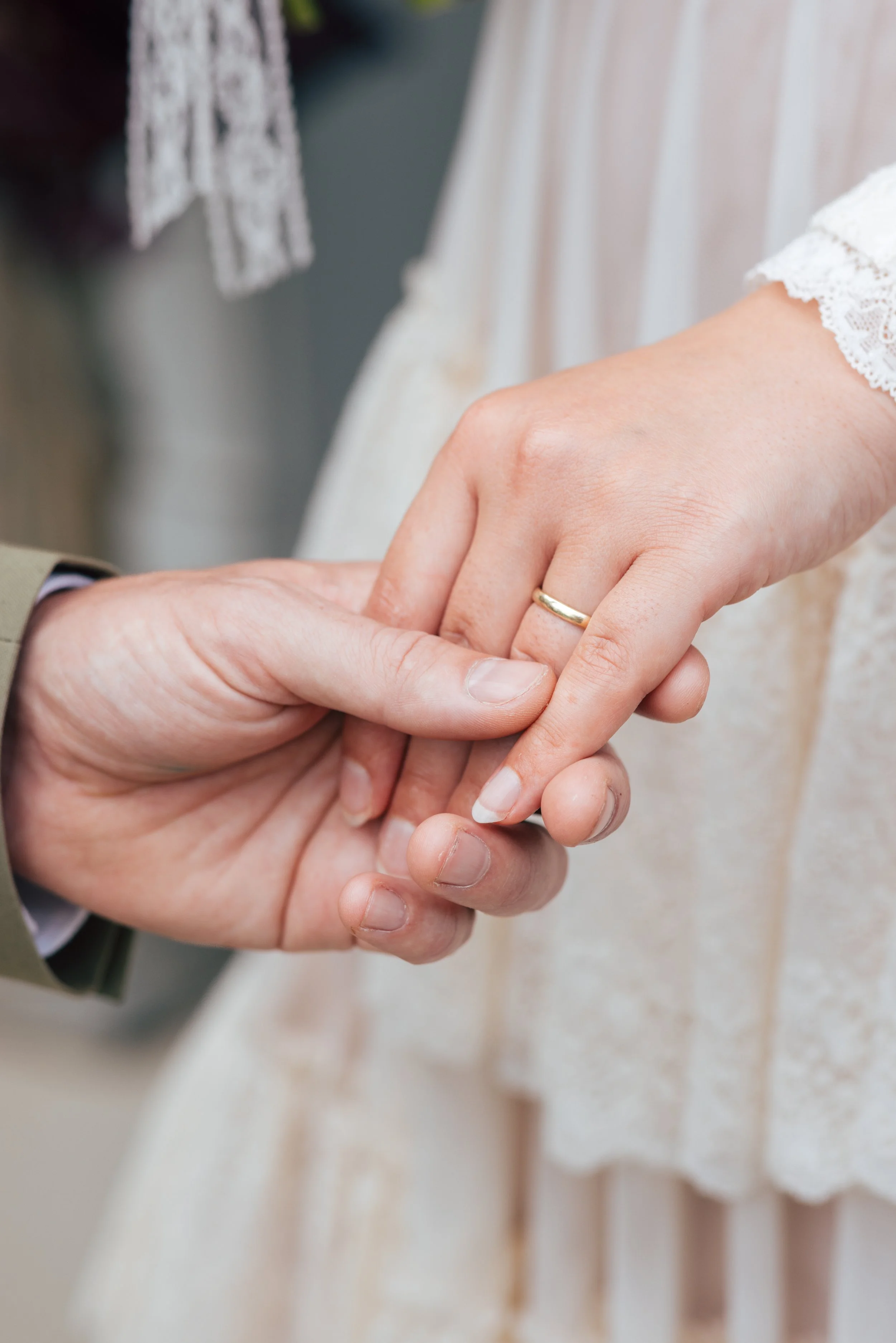 Close-up of a couple holding hands, with the woman wearing a wedding ring and a lace sleeve, indicating a wedding or engagement.