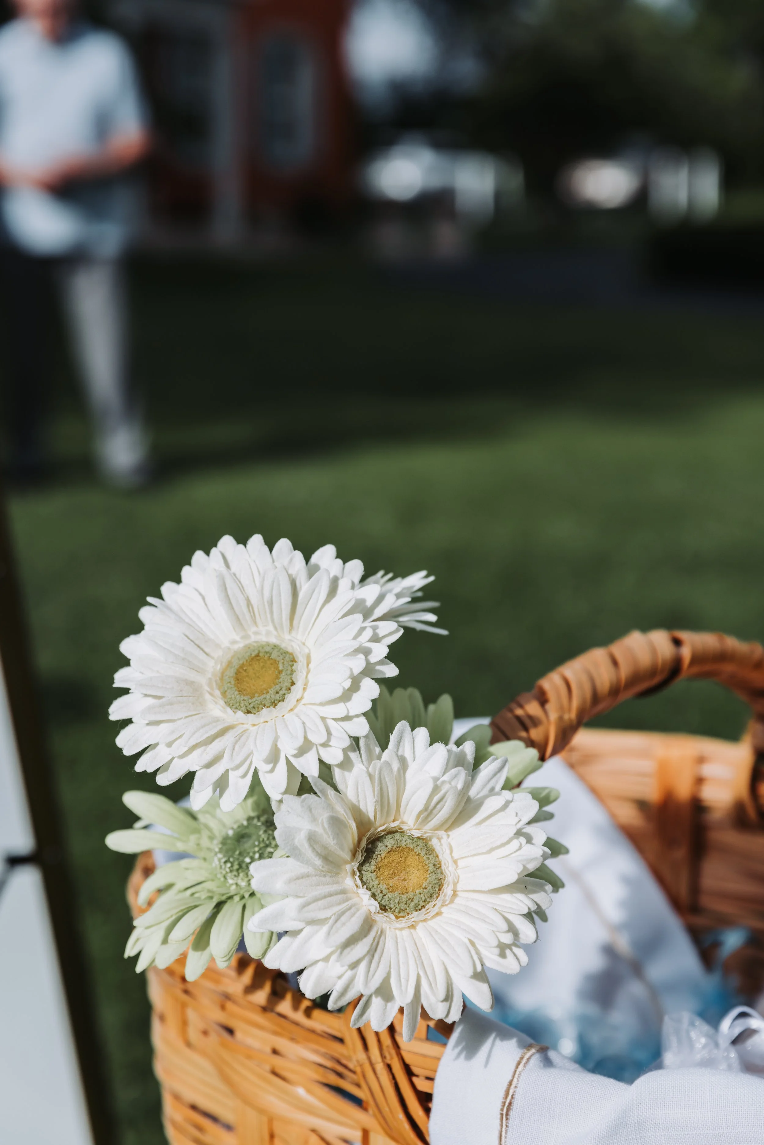 A wicker basket with white daisies and greenery on a table outdoors, with a blurred person in the background.