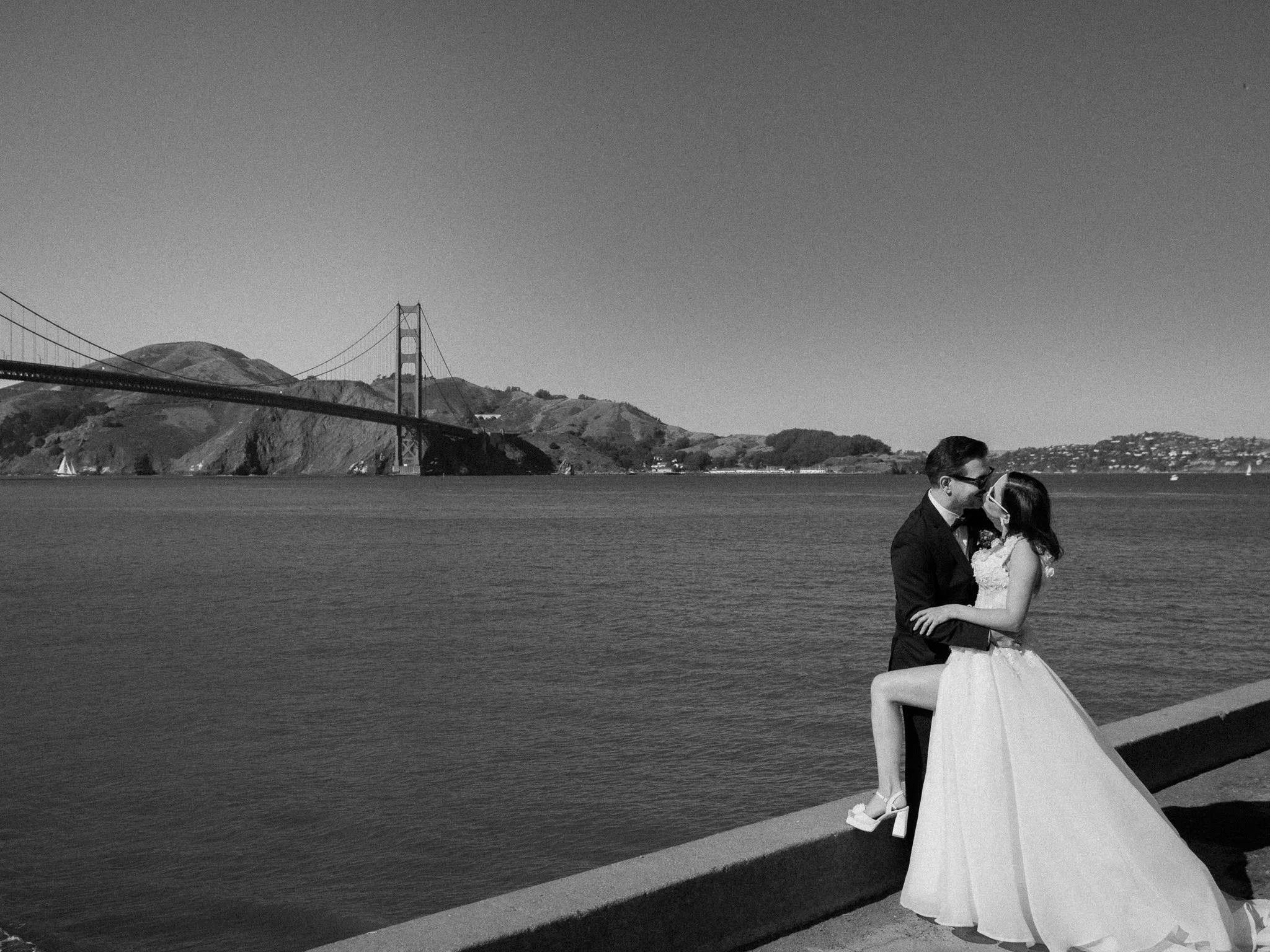 A black and white photo of a couple in wedding attire kissing near a body of water with the Golden Gate Bridge and hills in the background.