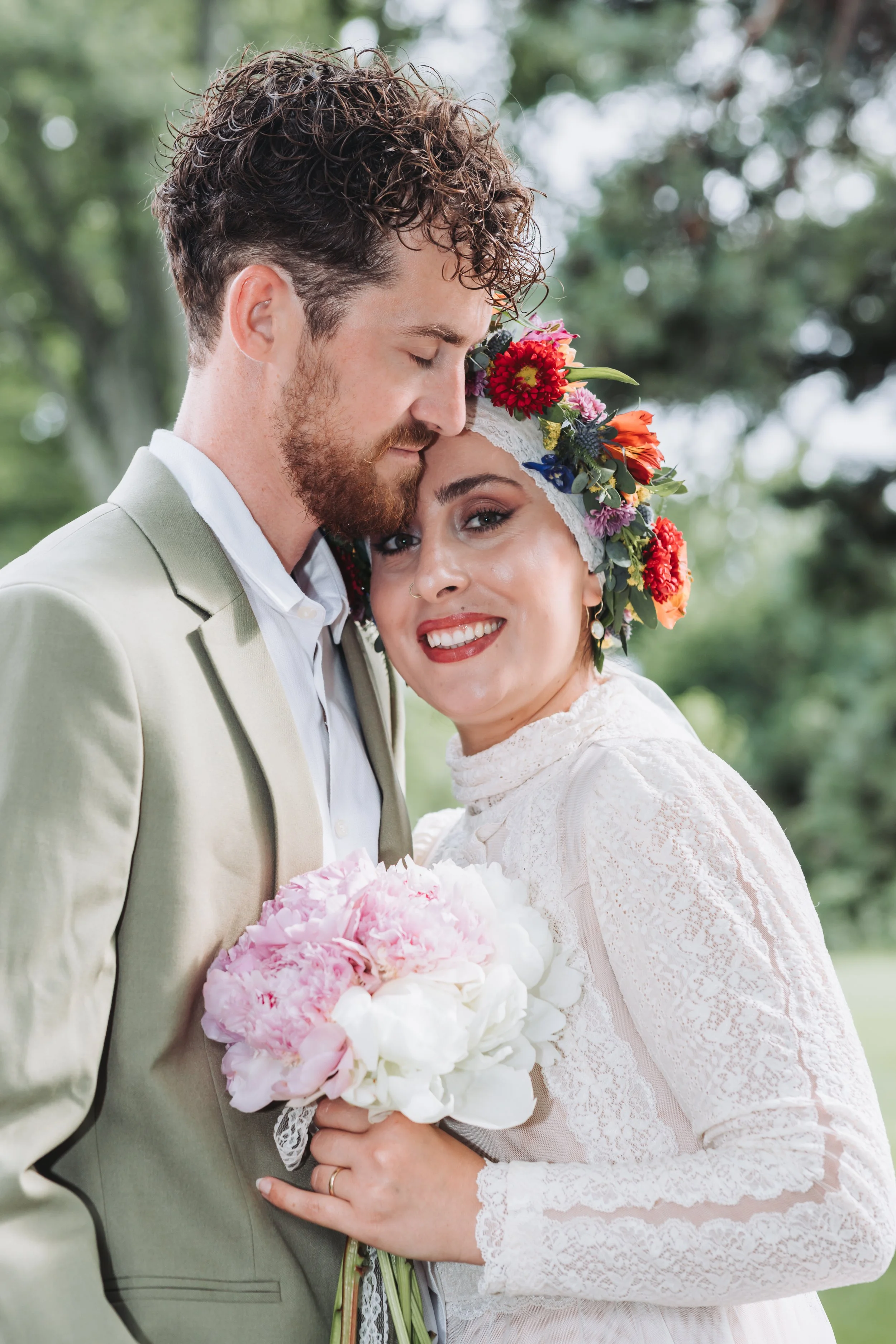 A newlywed couple poses outdoors, with the groom tenderly resting his forehead on the bride's head. The bride wears a white lace dress and a colorful floral headpiece, holding a bouquet of pink and white flowers. The groom wears a beige suit with a w