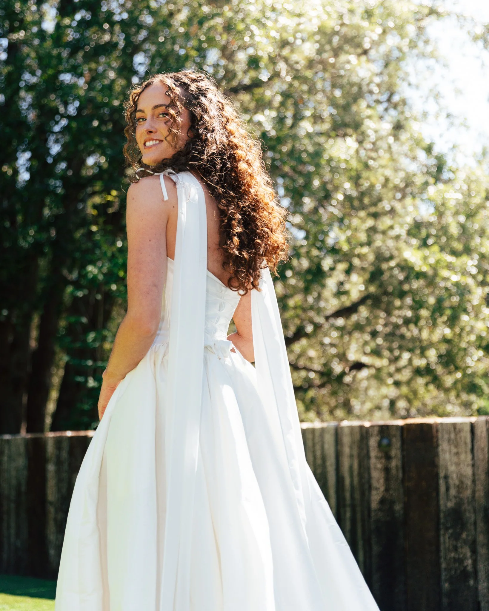 A woman in a white dress standing outdoors in sunlight, with lush green trees in the background.