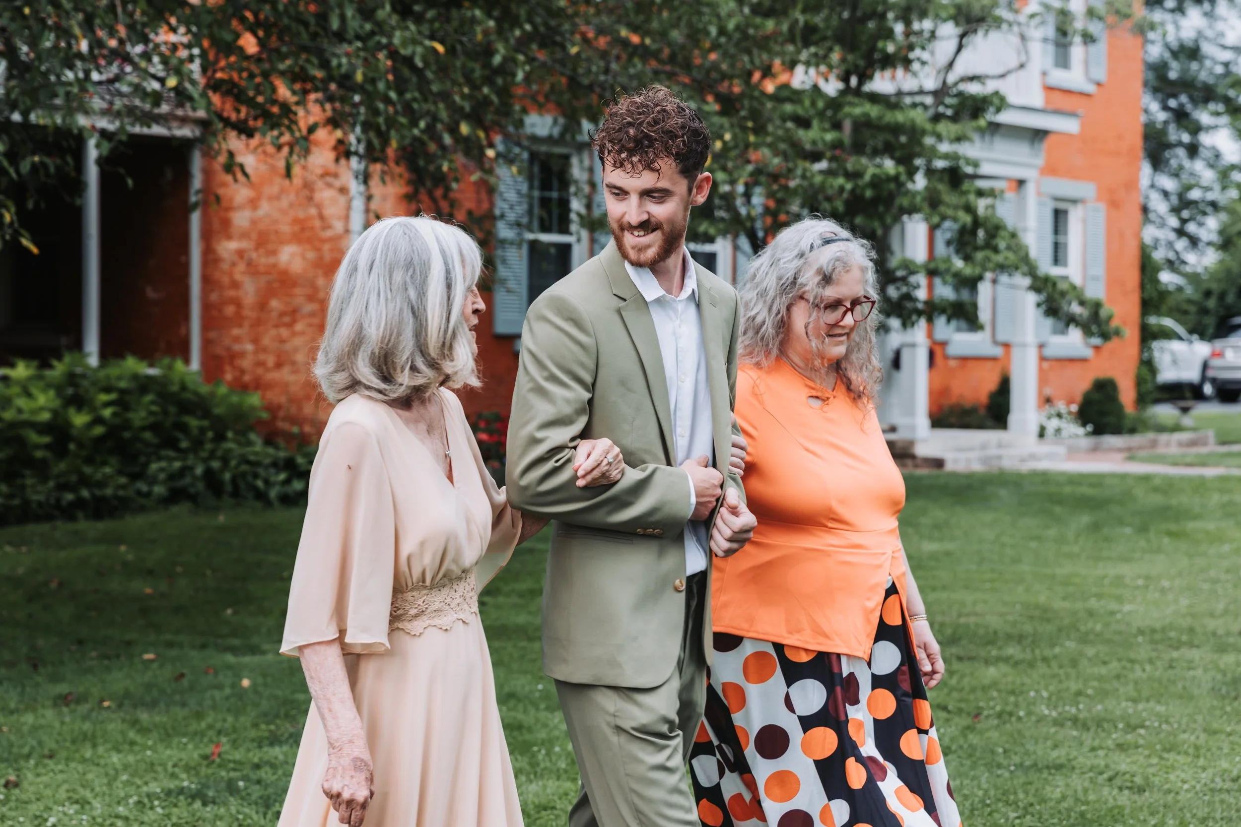 A young man in a light green suit walks arm-in-arm with two elderly women outdoors, with a brick house and greenery in the background.