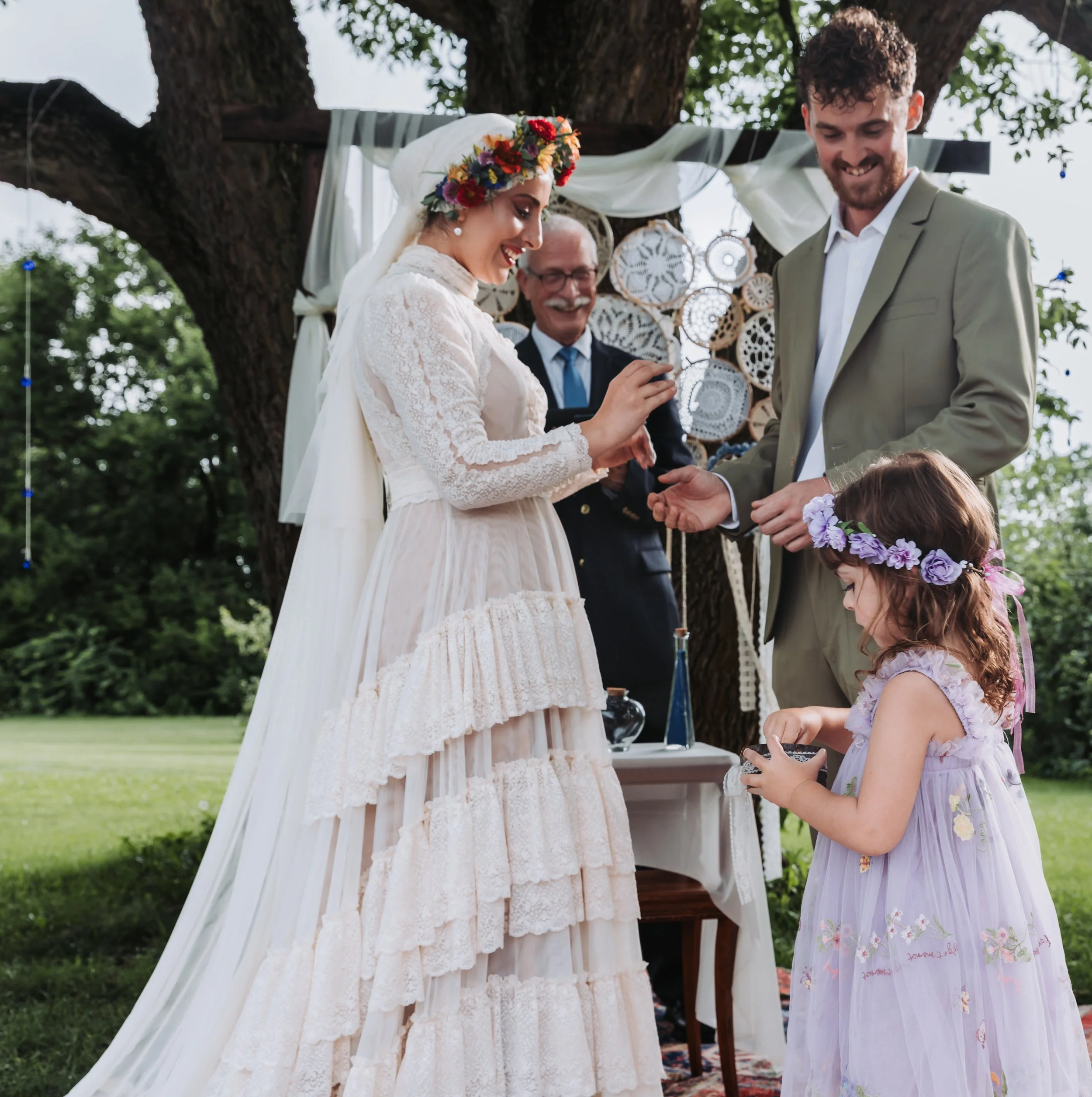 A woman in a vintage wedding dress wearing a flower crown exchanges rings with a man in a light green suit during an outdoor wedding ceremony, with a small girl in a purple dress and floral headband nearby, and an officiant standing behind them.