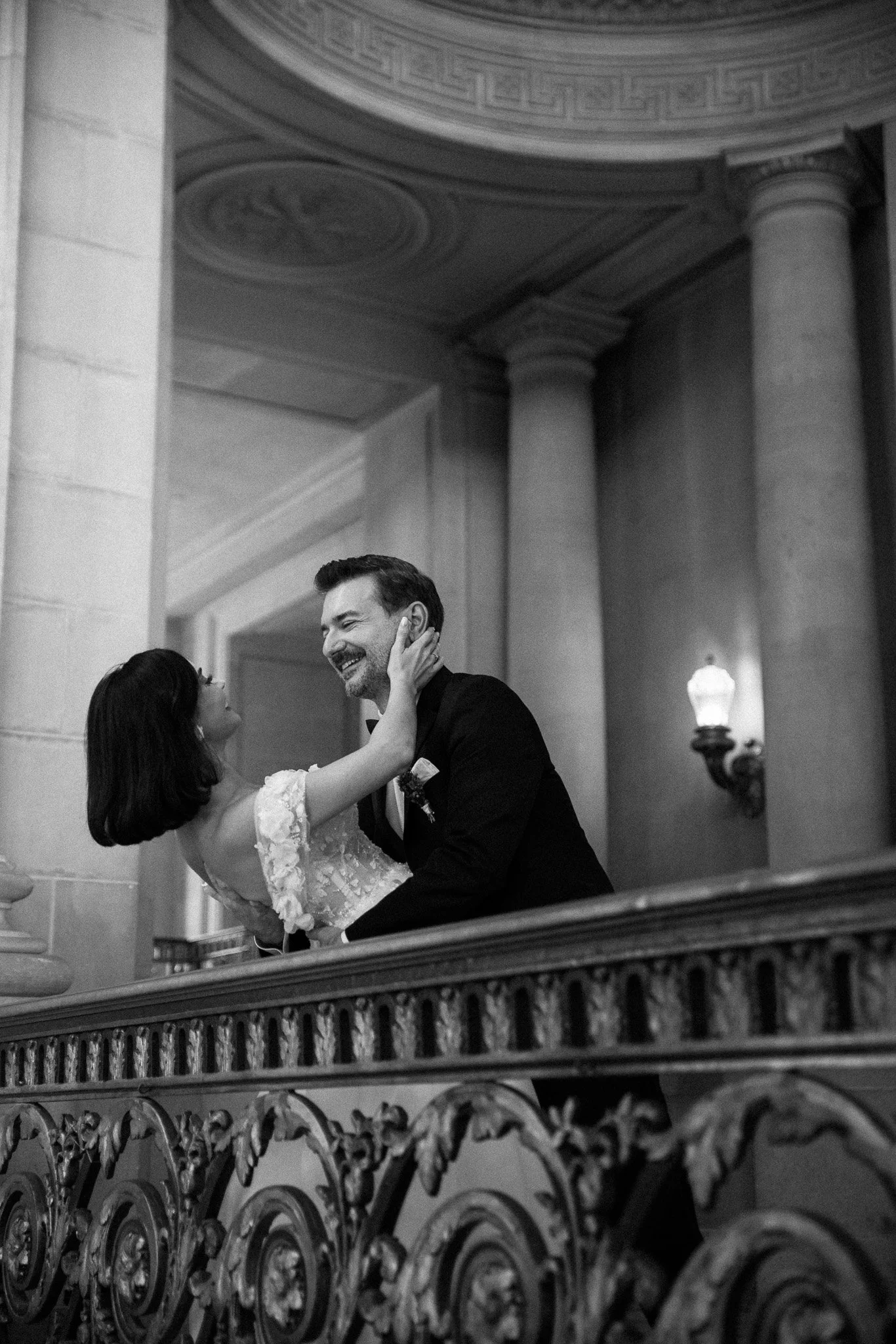 A black and white photo of a man in a tuxedo dancing with a woman in a lace dress in an elegant hall, with ornate ironwork and classical columns in the background.