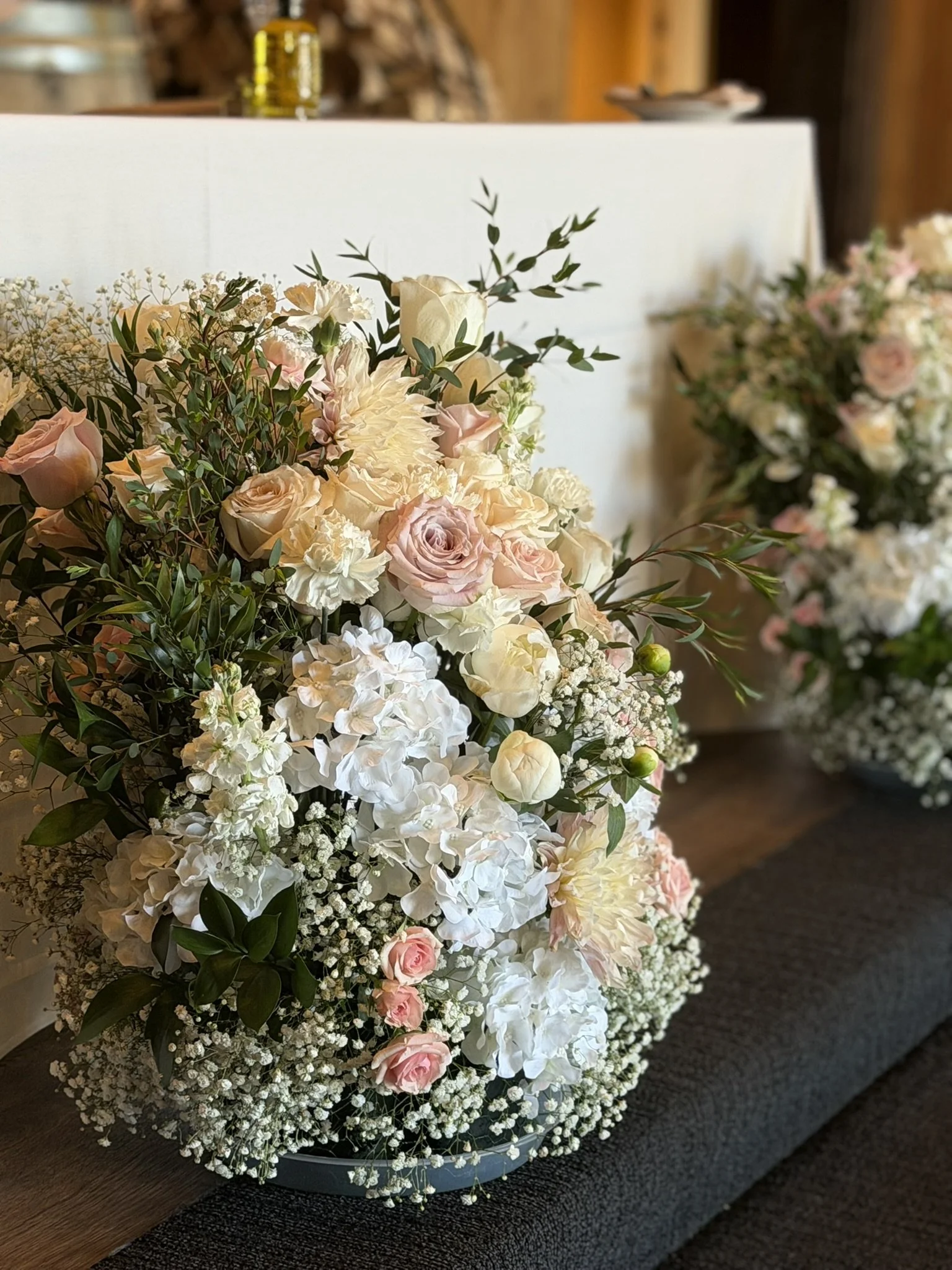A bouquet of pink and white flowers, including roses, hydrangeas, and baby's breath, arranged on a wooden table against a blurred background.