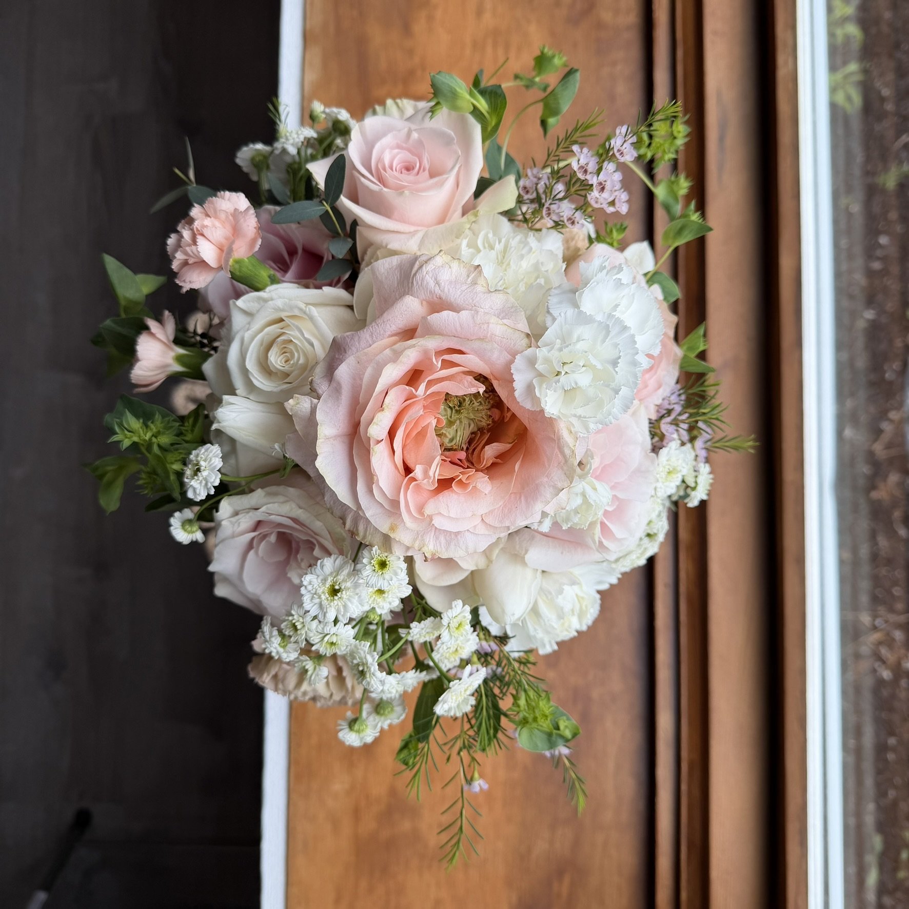 A bouquet of pink and white roses, carnations, and baby’s breath flowers on a wooden surface near a window.