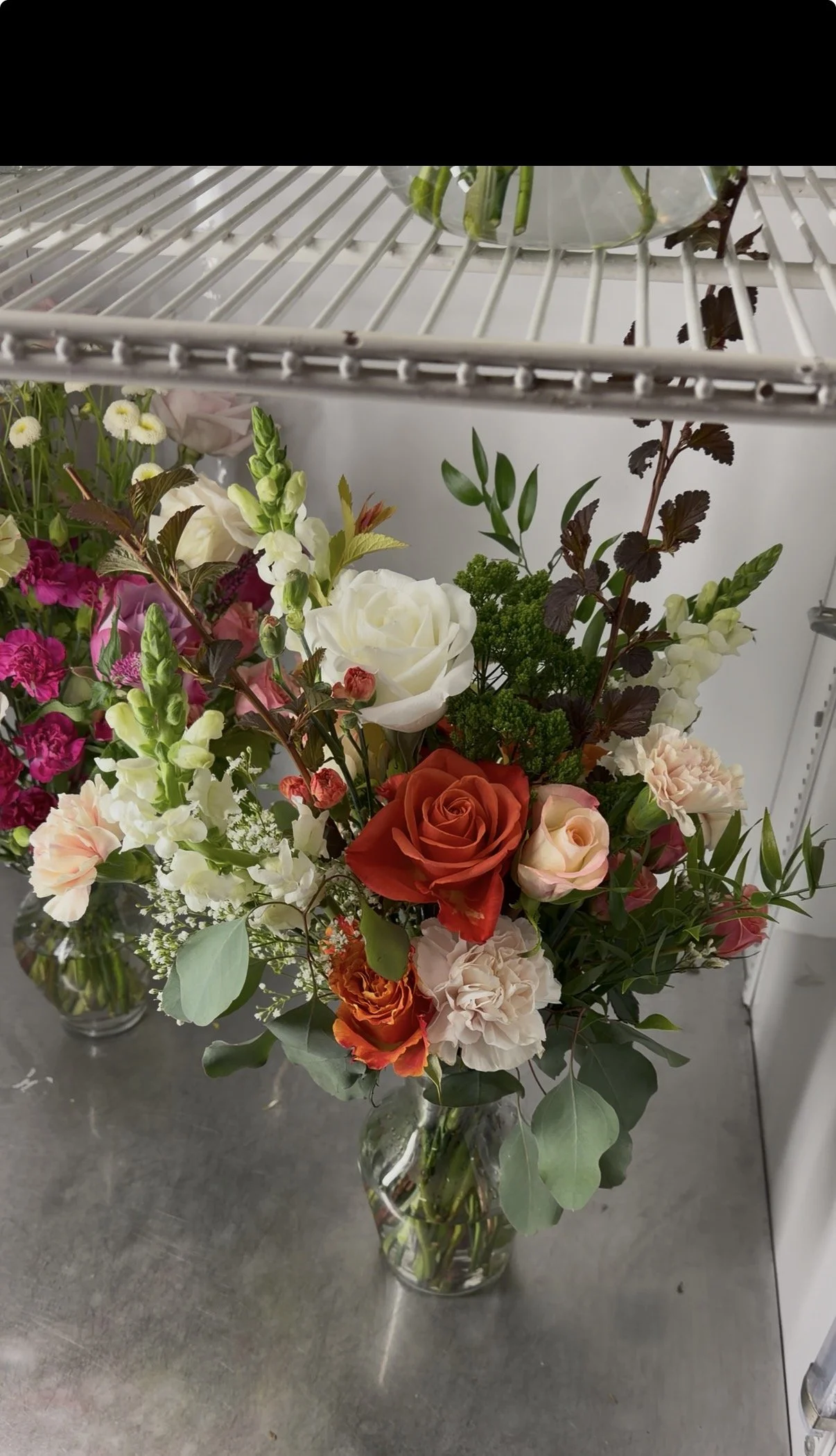 A bouquet of mixed flowers including roses, carnations, and greenery arranged in a glass vase, placed on a gray surface.
