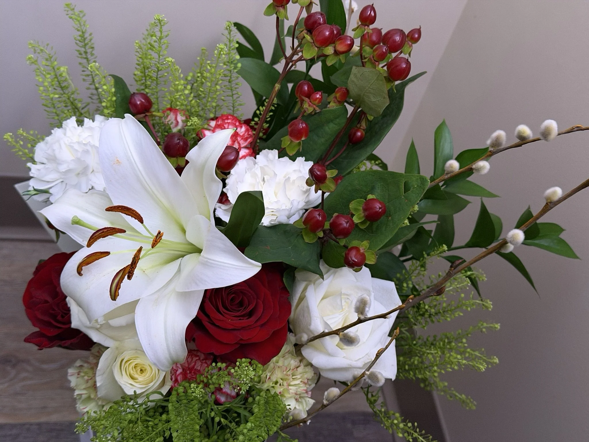 A colorful floral arrangement featuring white lilies, red roses, white carnations, green foliage, and berries.
