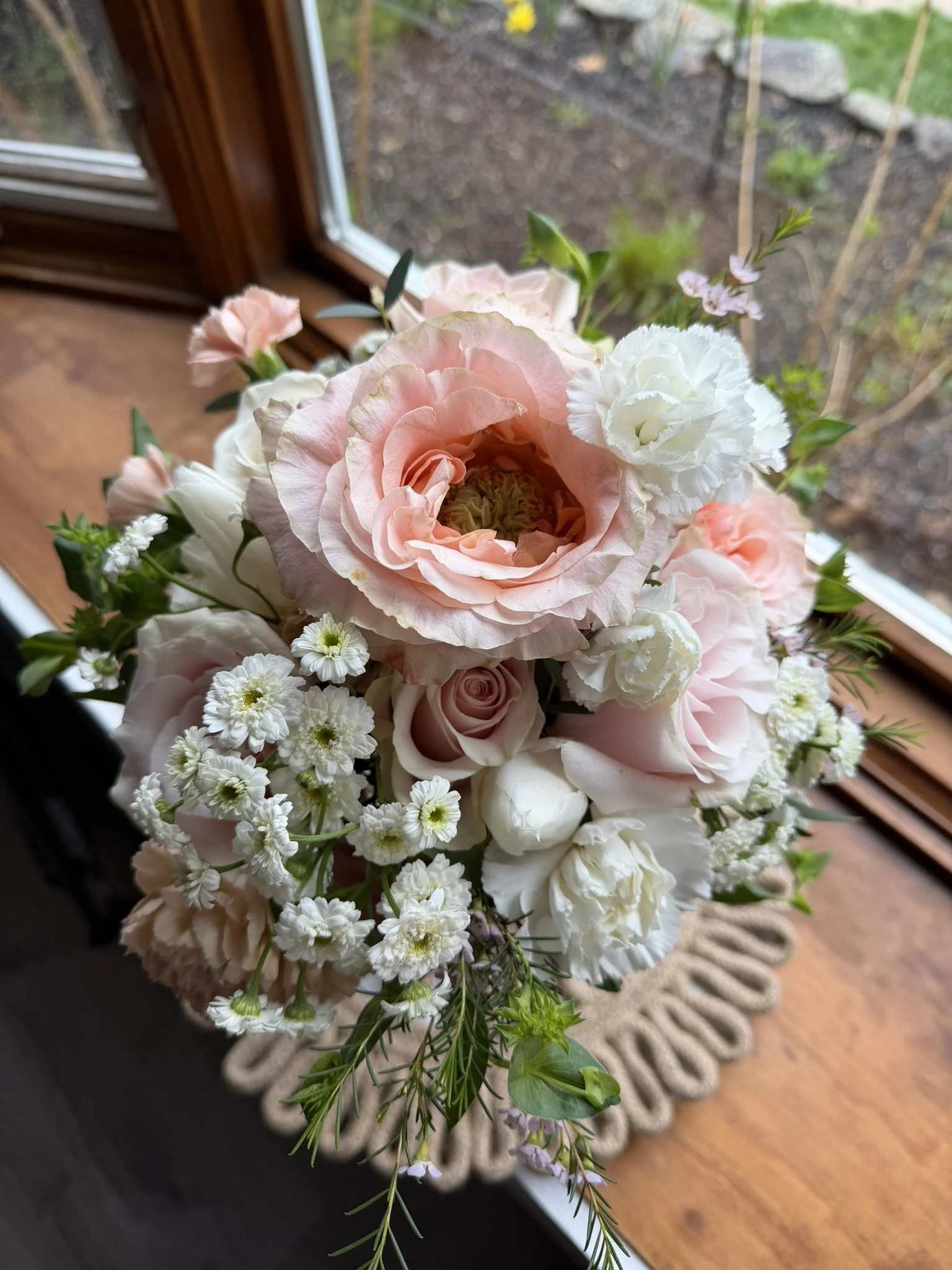 A bouquet of pink and white flowers with greenery on a wooden surface near a window showing a garden outside.