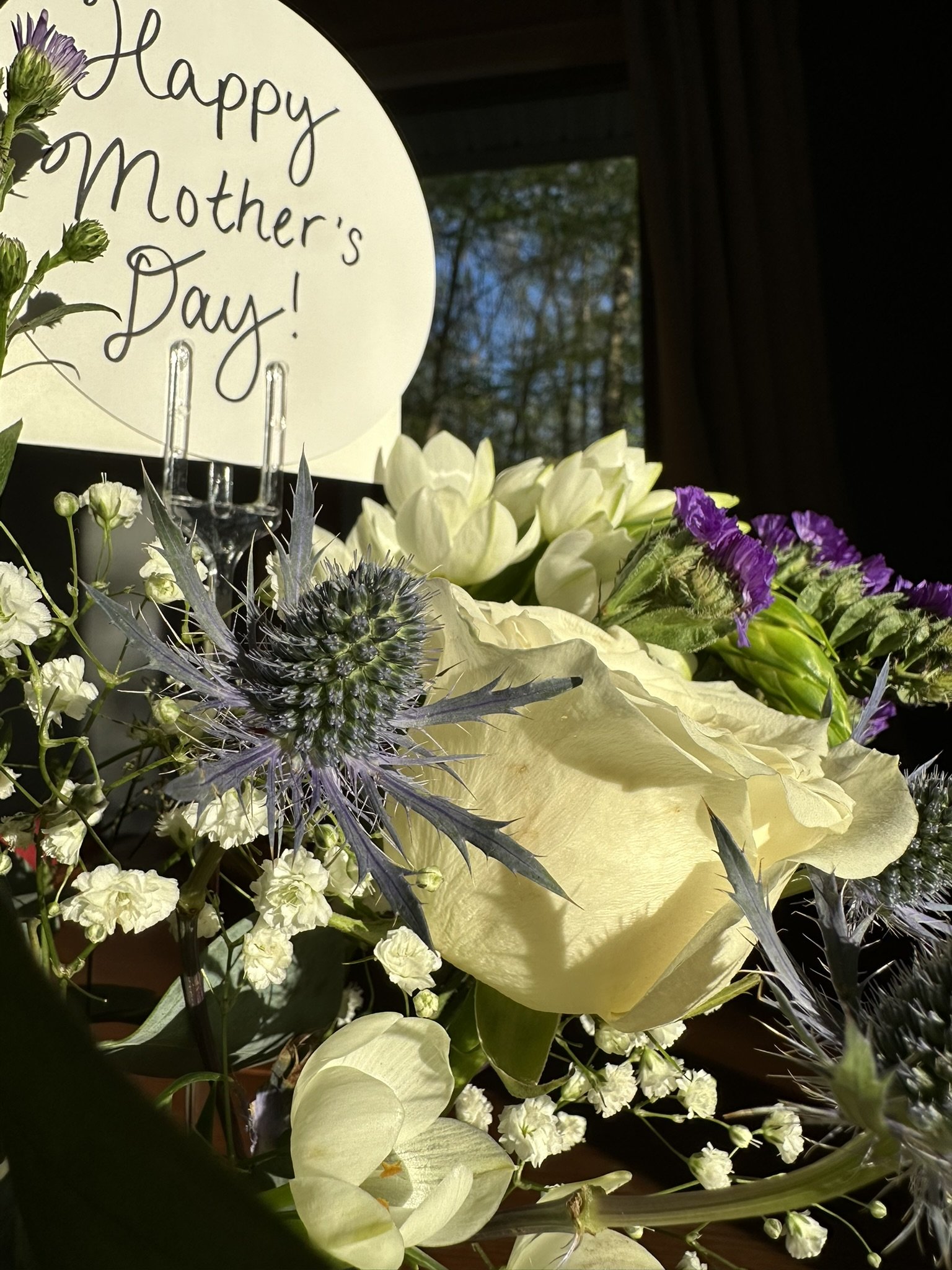 A floral arrangement with white, purple, and green flowers next to a sign that says 'Happy Mother's Day!'