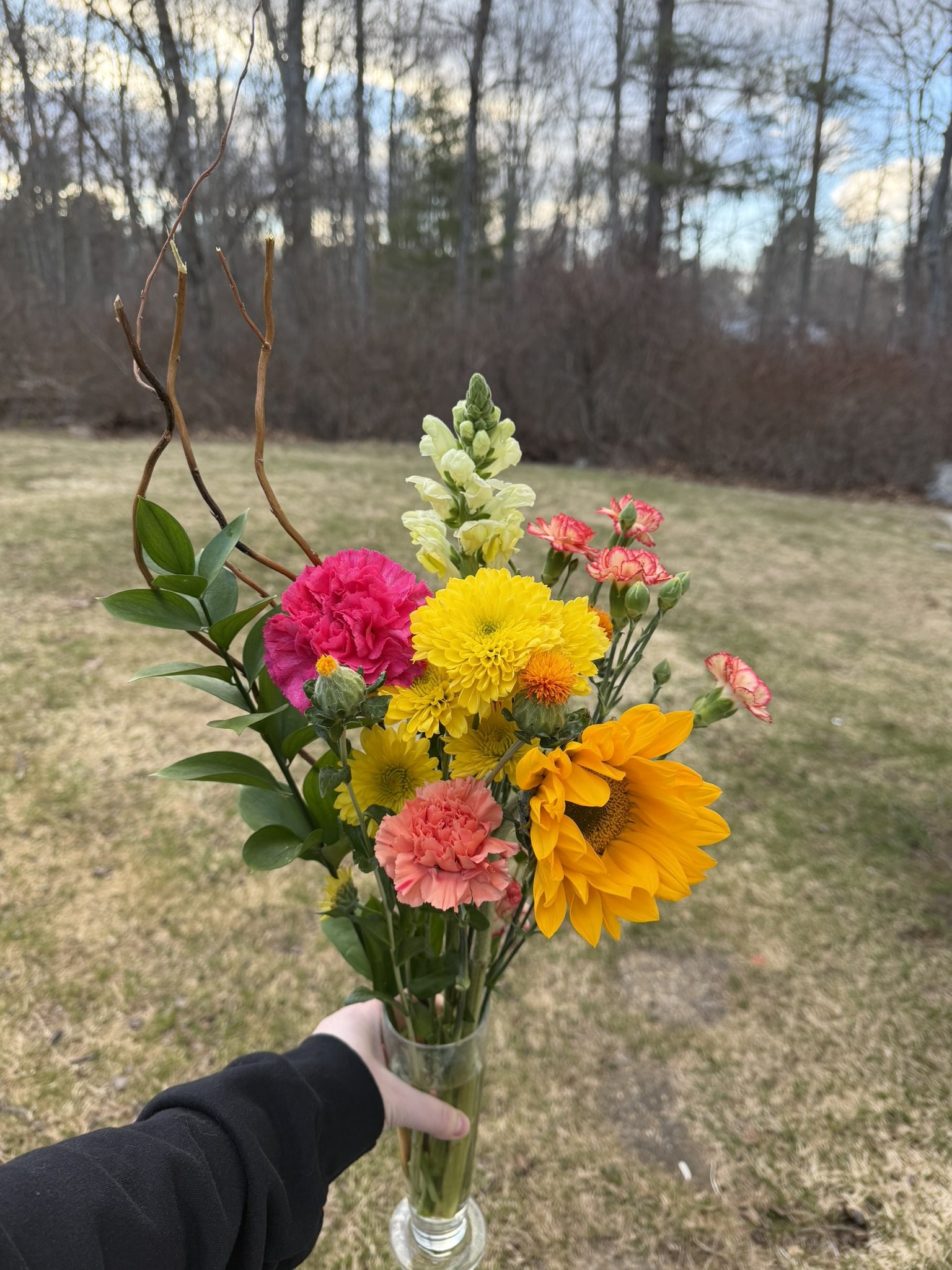 A person holding a glass vase filled with a colorful assortment of fresh flowers outdoors against a background of a grassy yard and trees.