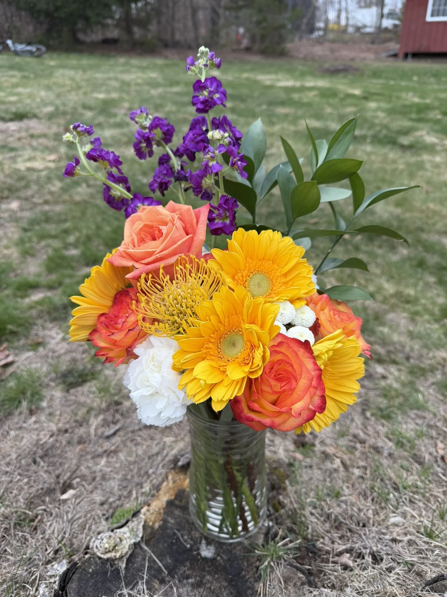 Colorful bouquet of flowers in a glass vase placed outdoors on the ground, with a grassy yard and trees in the background.