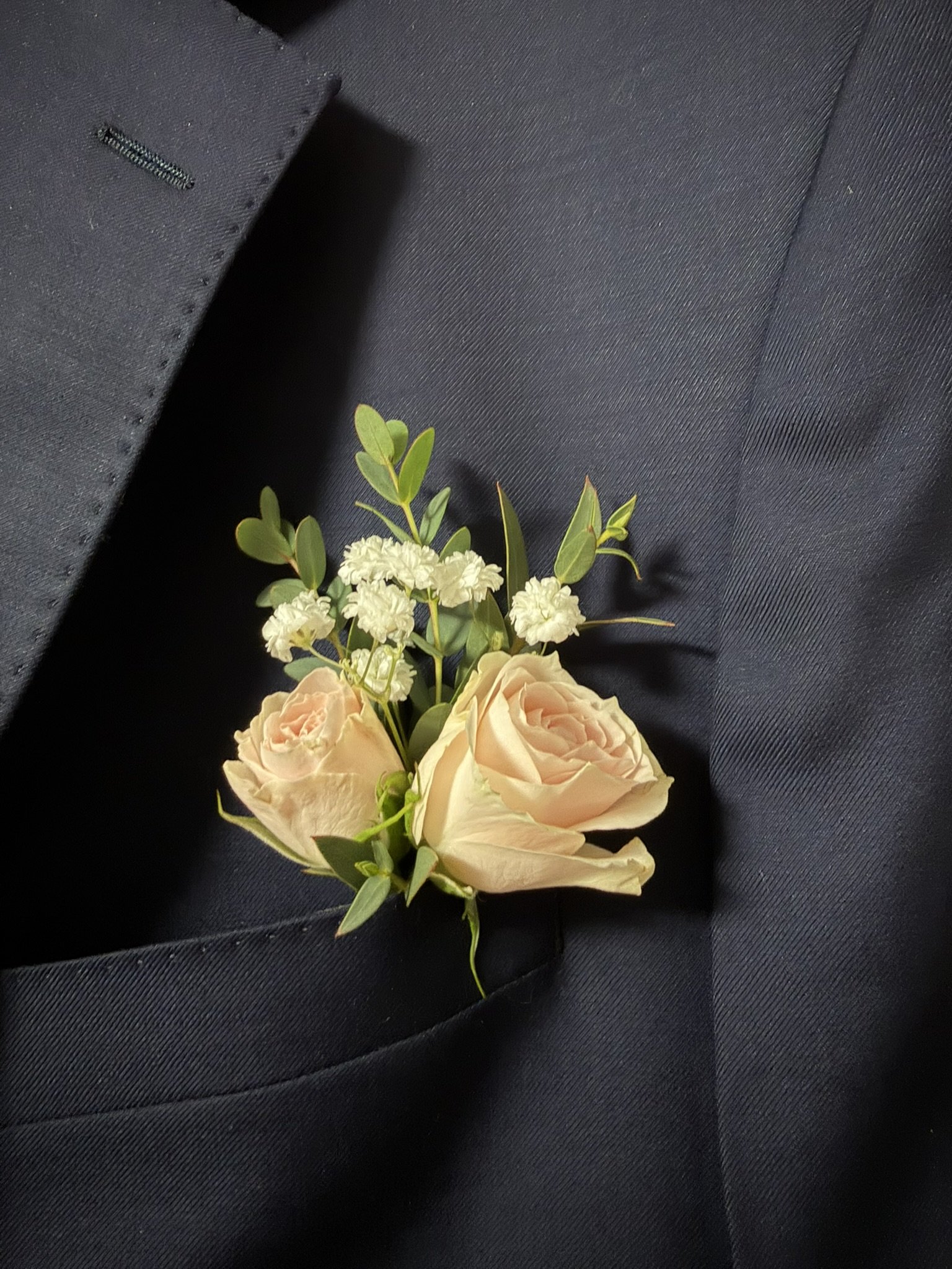 A boutonniere with light pink roses, white filler flowers, and green foliage attached to a black suit lapel.