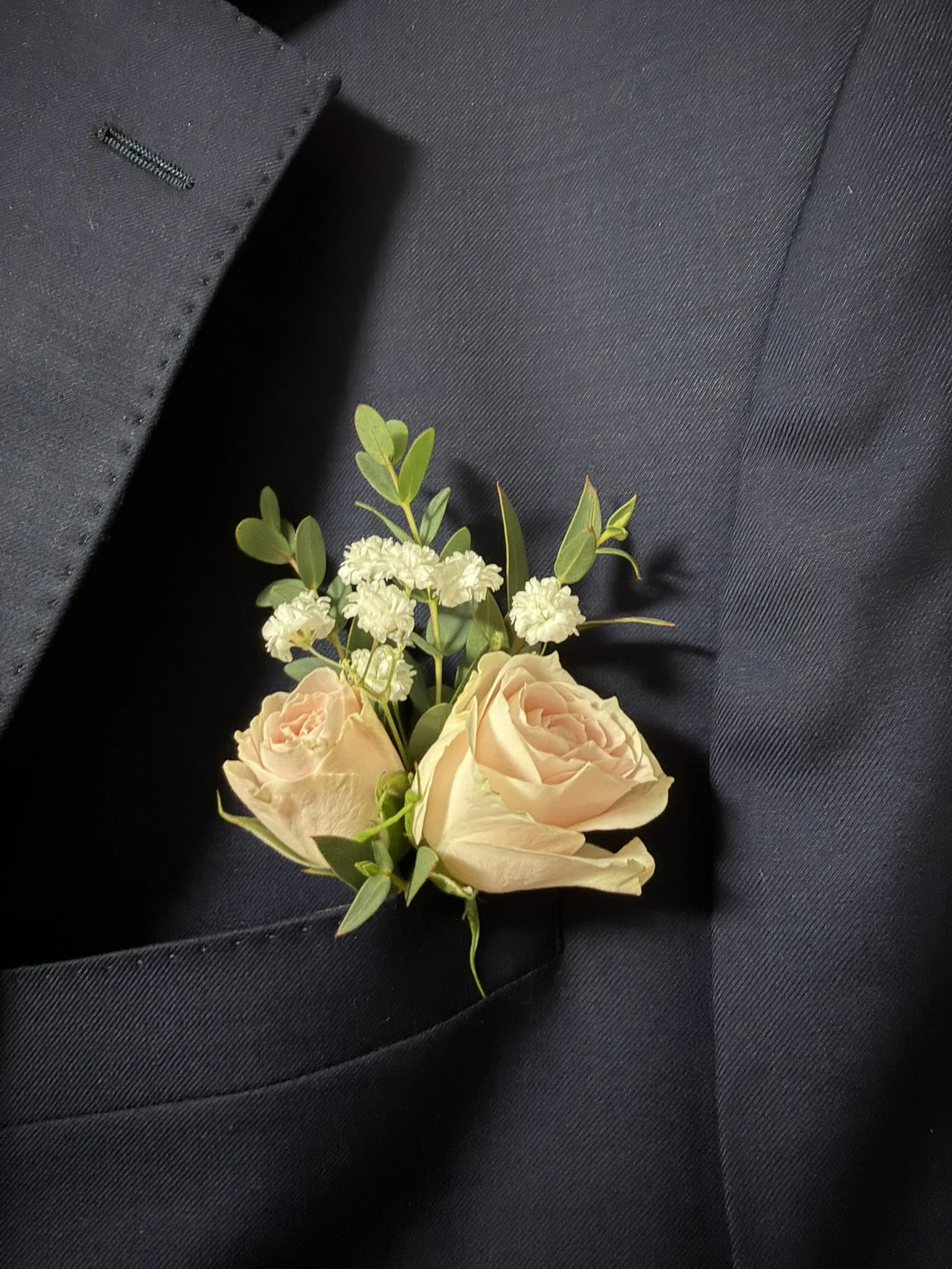 Close-up of a boutonniere with cream roses, white small flowers, and green leaves, pinned to a dark suit fabric.