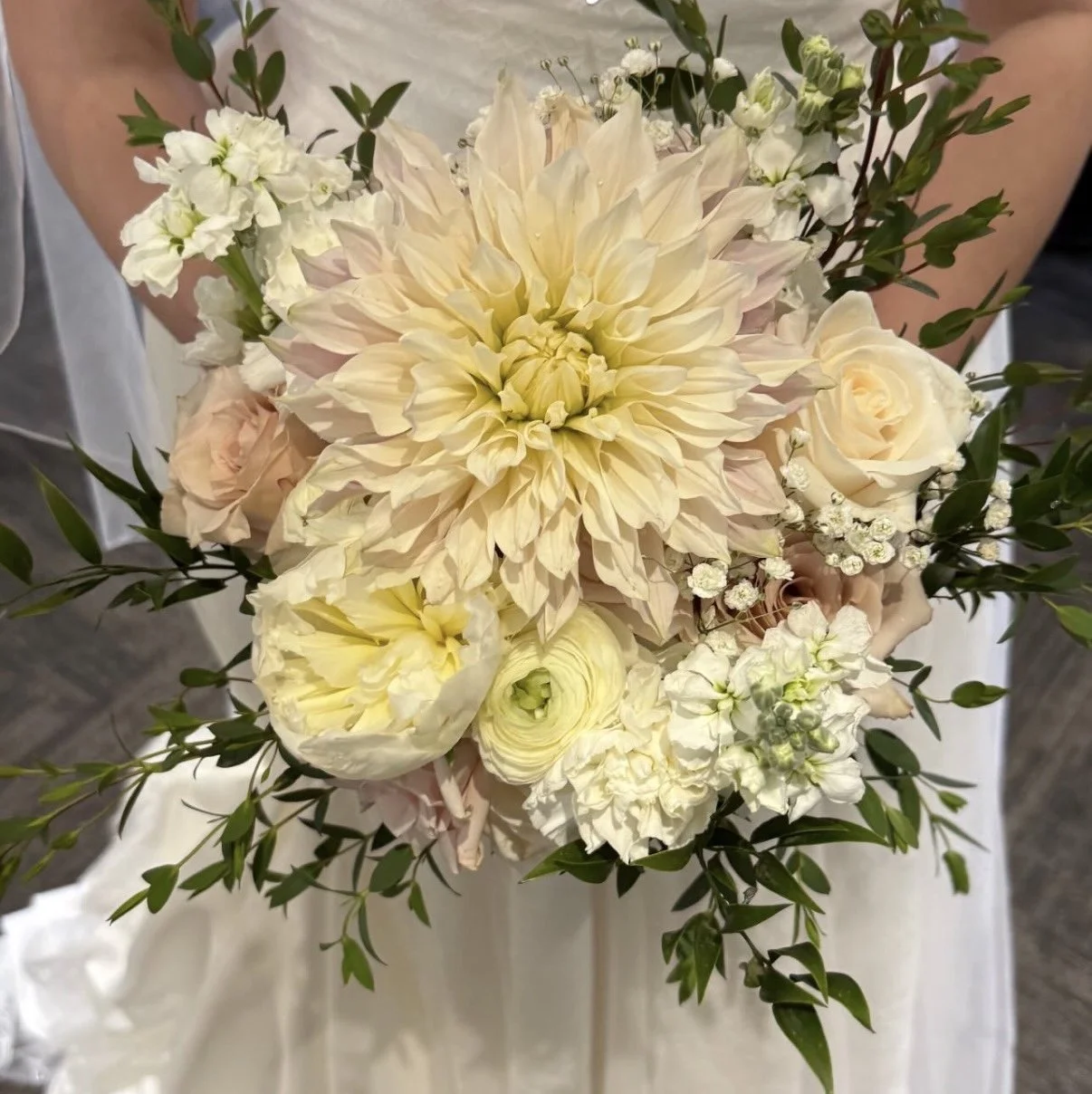 A person holding a bouquet of ivory, white, and pale pink flowers including dahlias, roses, and small filler flowers with green foliage.