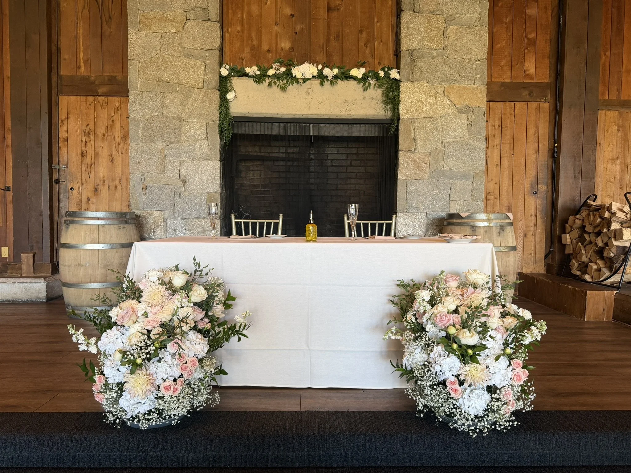 A decorated indoor setting with a fireplace, floral arrangements on a table and on the floor, with wine glasses, a bottle, and candles on the table.