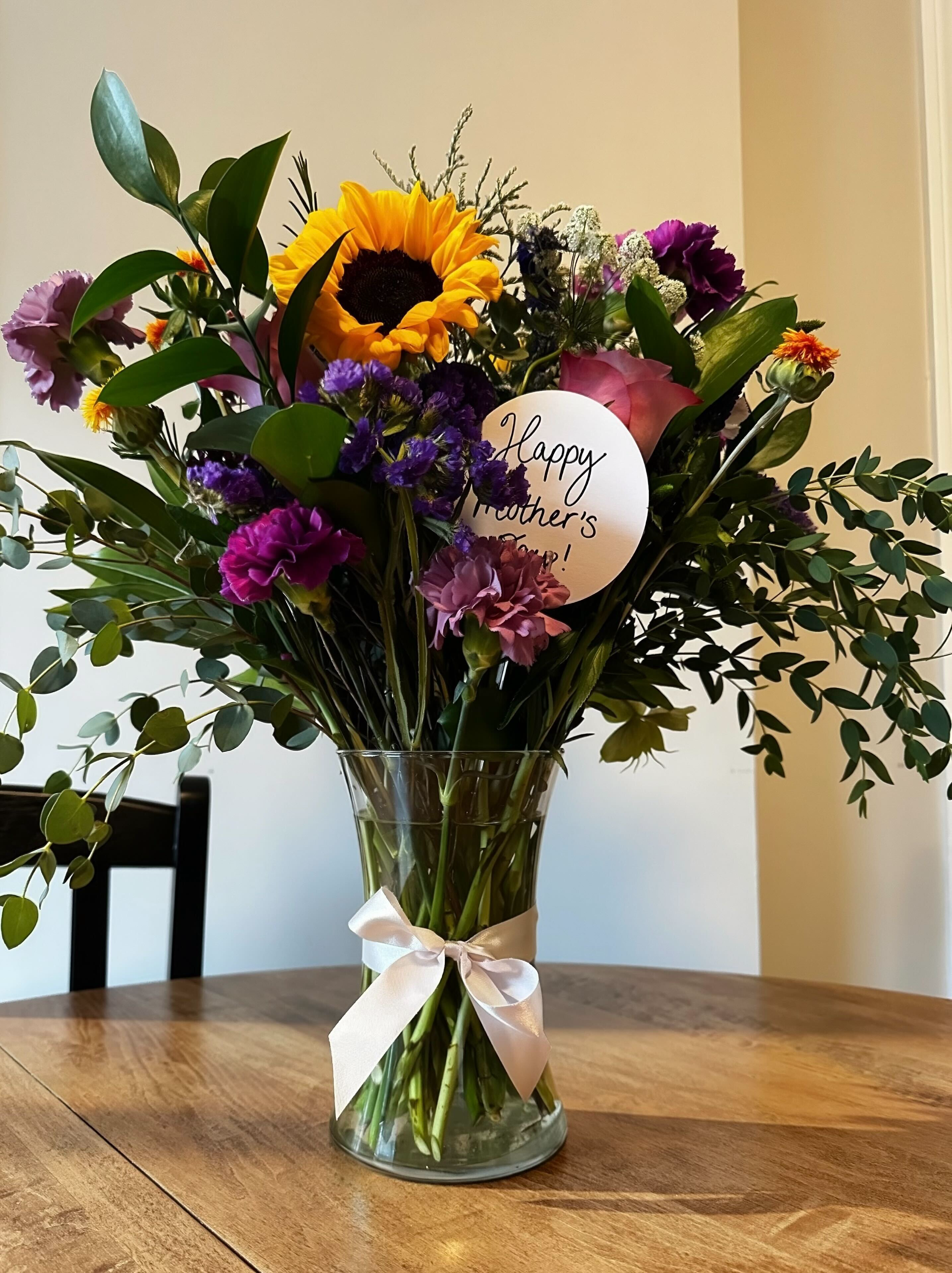 A bouquet of mixed flowers including sunflowers, pink roses, purple carnations, and small white filler flowers in a glass vase with a cream-colored ribbon, placed on a wooden table.
