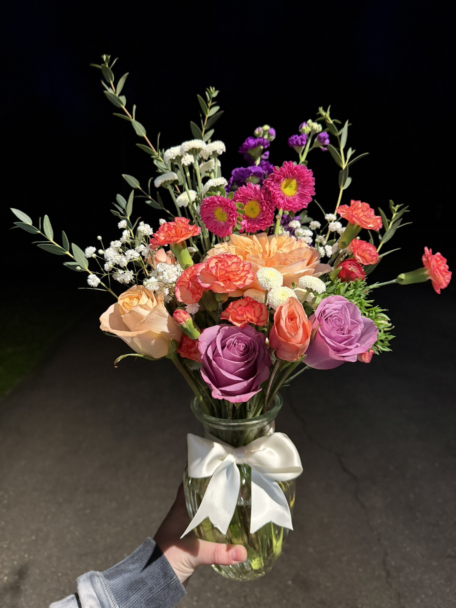 Hand holding a glass vase with a colorful mixed flower bouquet against a dark background.