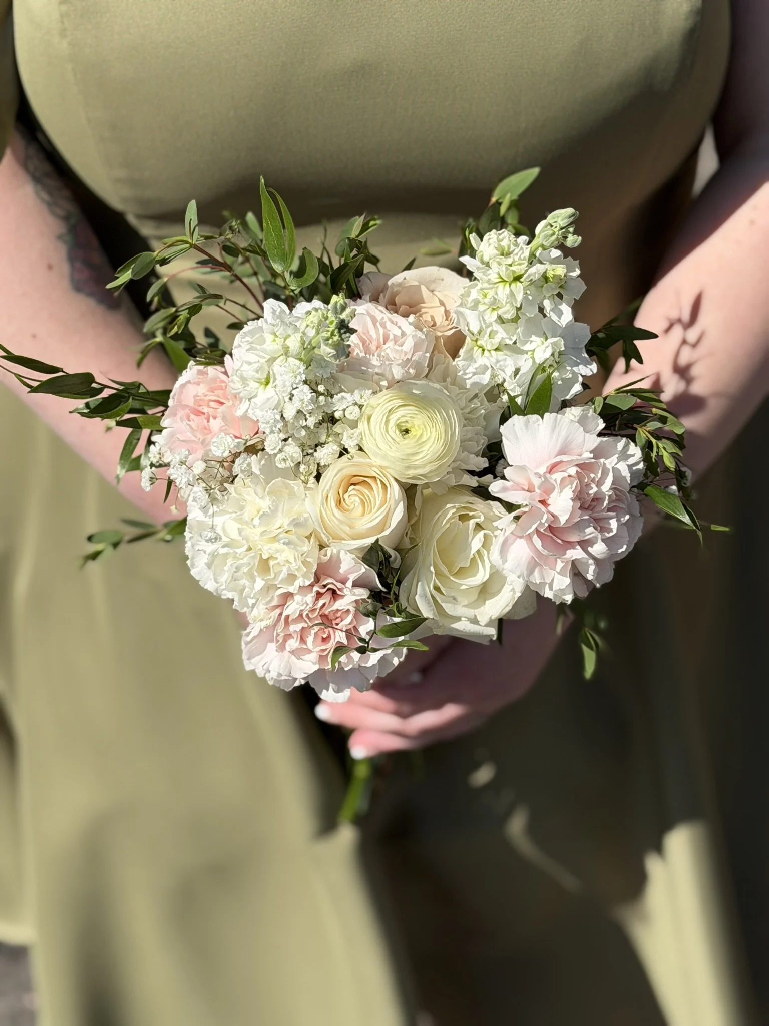 A person holding a bouquet of white and pale pink flowers with green foliage, wearing a green dress with a tattoo visible on their arm.