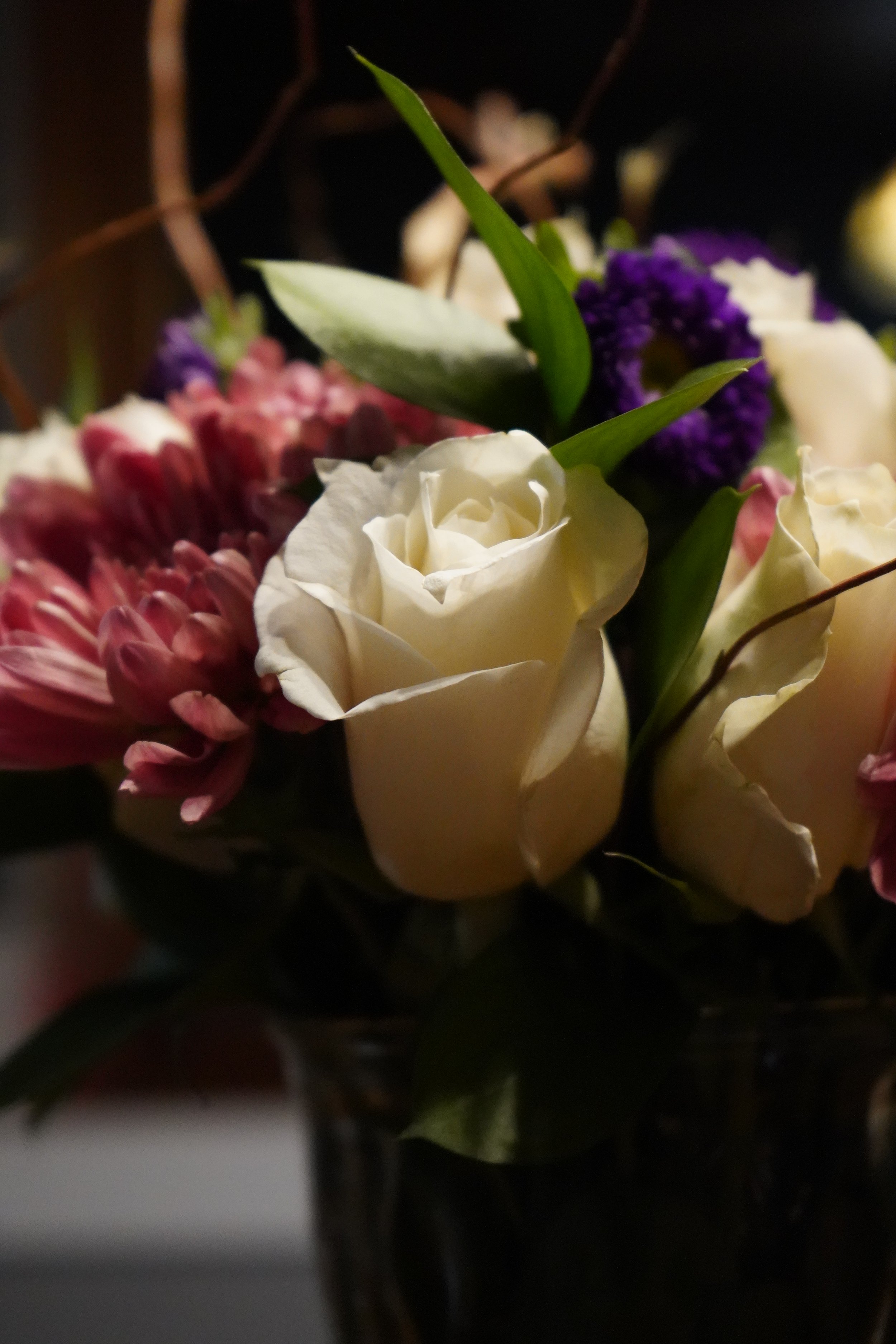 A bouquet of various flowers, including white roses, purple and pink flowers, with green leaves, in a dark vase.