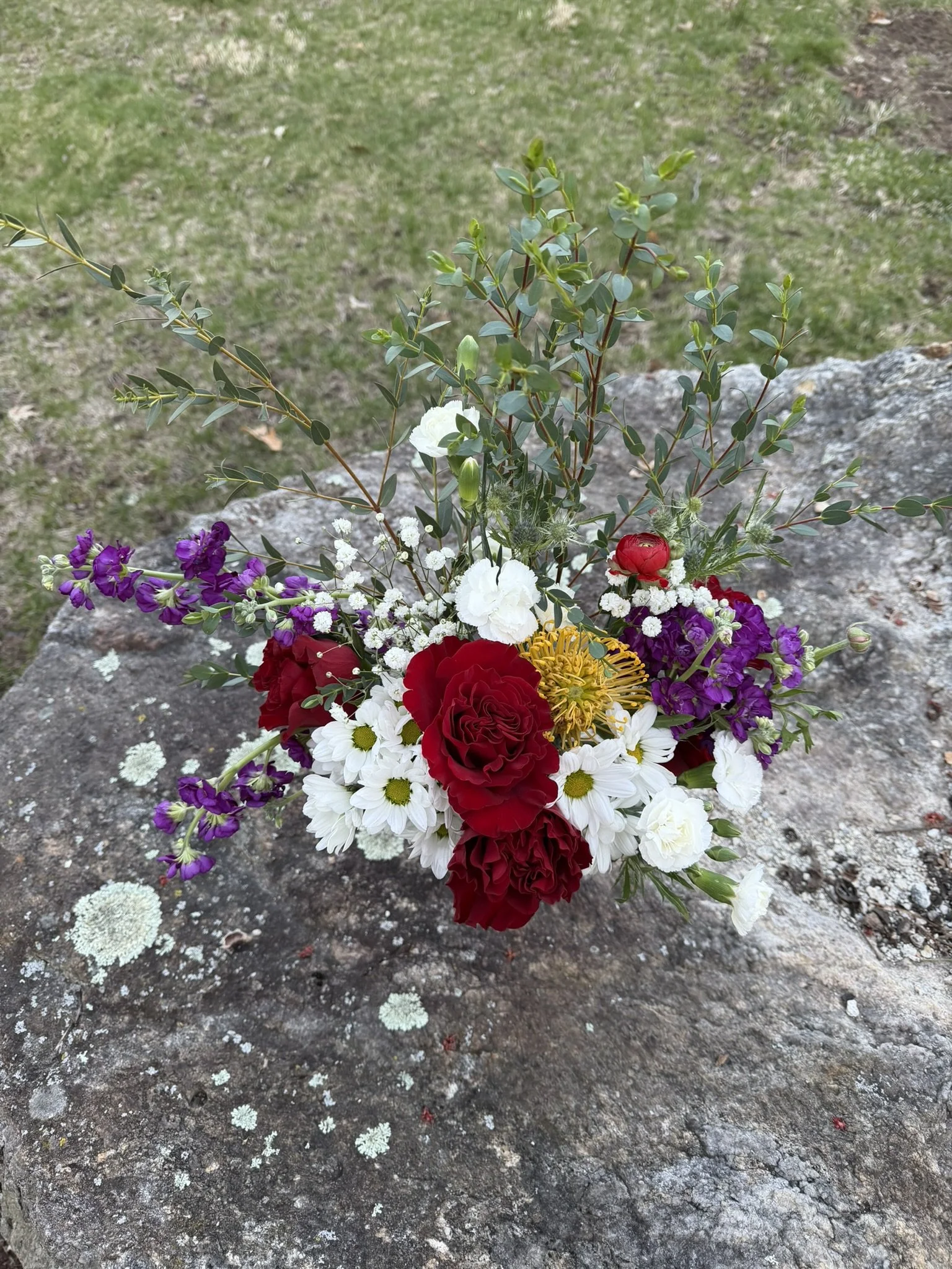 A bouquet of various flowers including red roses, white daisies, purple flowers, and small white fillers, placed on a large rock outdoors.