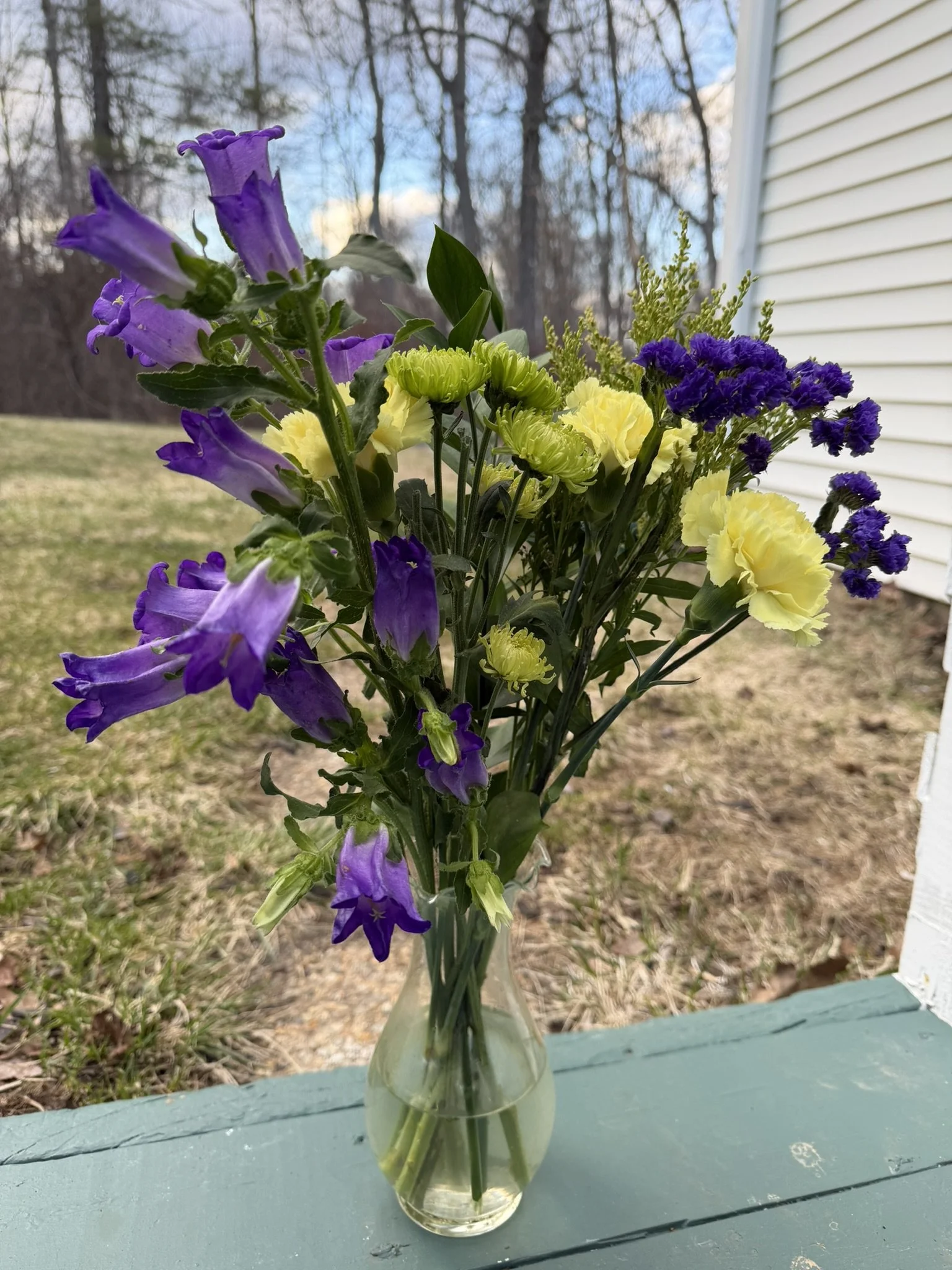 A glass vase filled with a bouquet of purple, yellow, and purple flowers on a light blue wooden surface outdoors with a background of trees and a house siding.