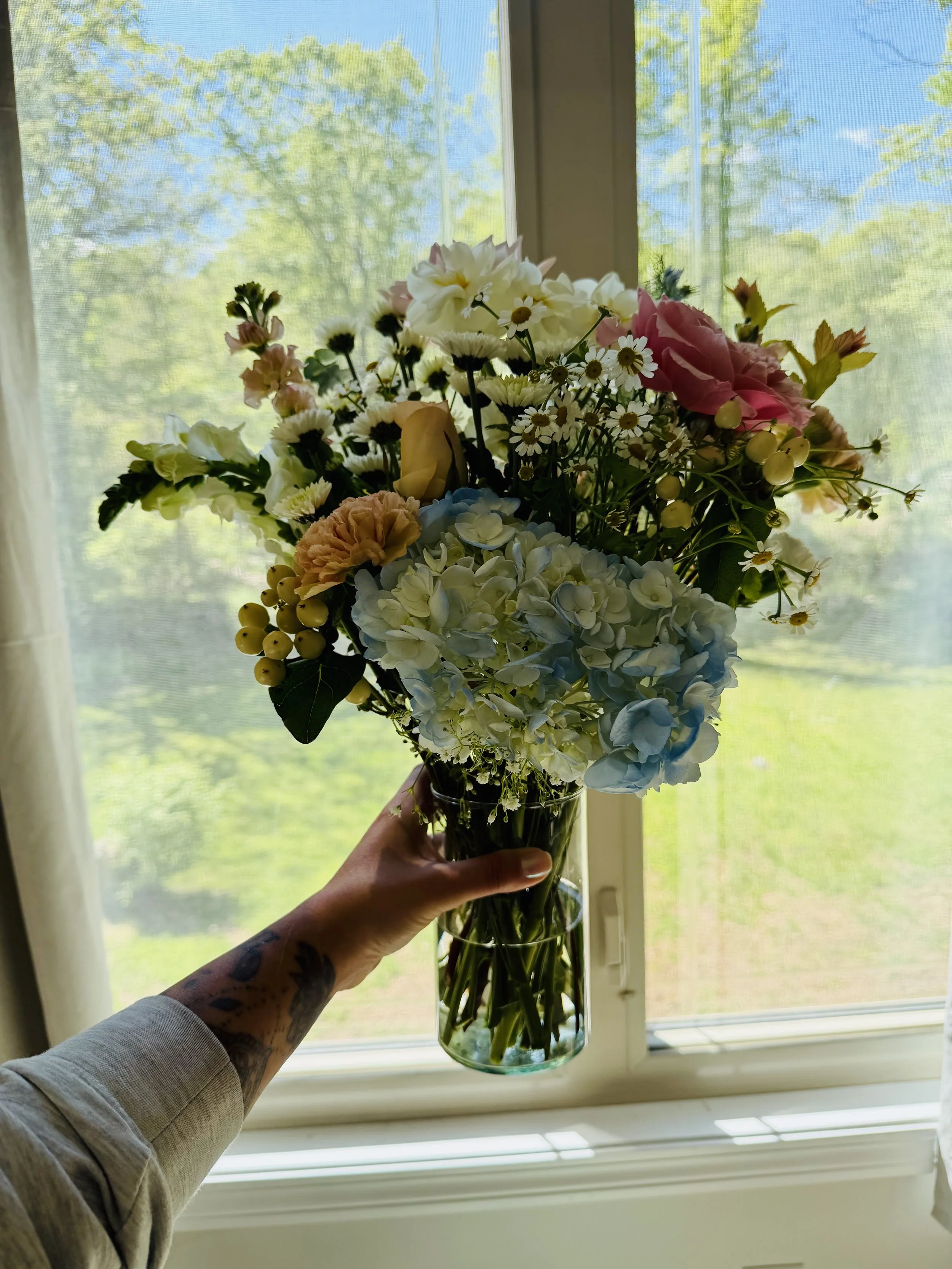 Person holding a bouquet of mixed flowers in front of a window with trees outside.