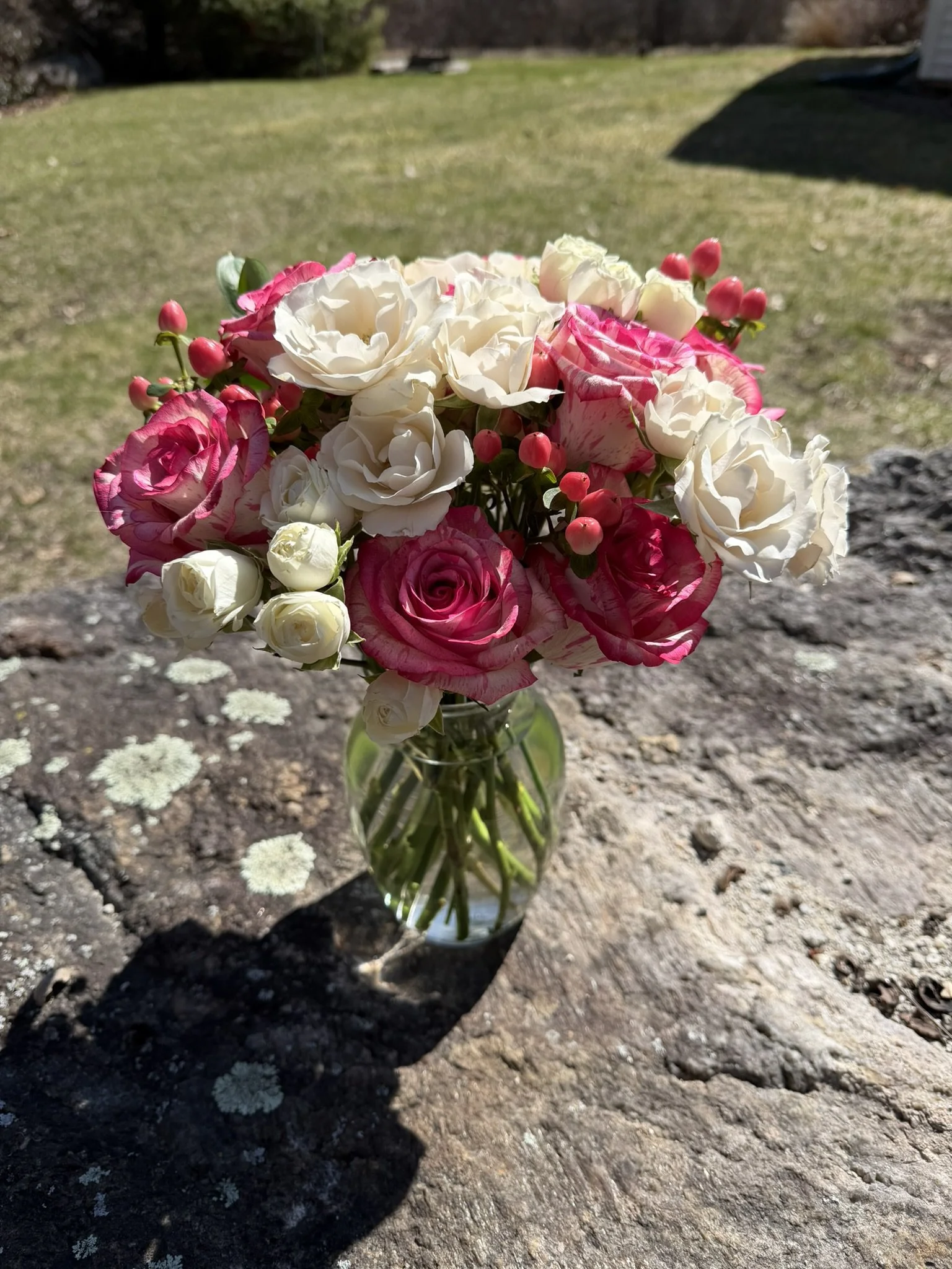A bouquet of pink and white roses in a glass vase on a stone surface outdoors with grass and trees in the background.