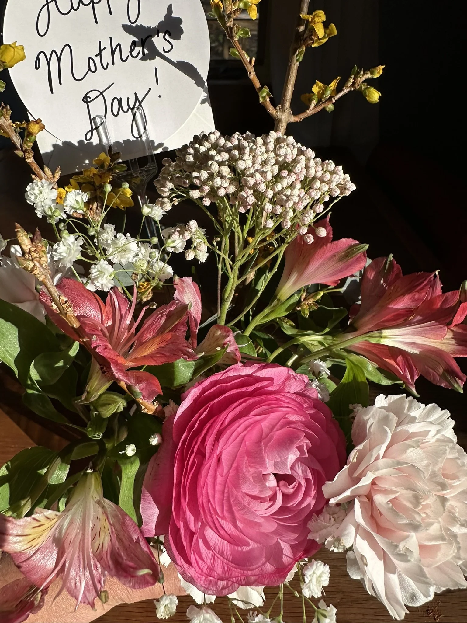 A floral arrangement with pink, white, and red flowers, small white buds, and branches, with a sign in the background that says "Happy Mother's Day!"