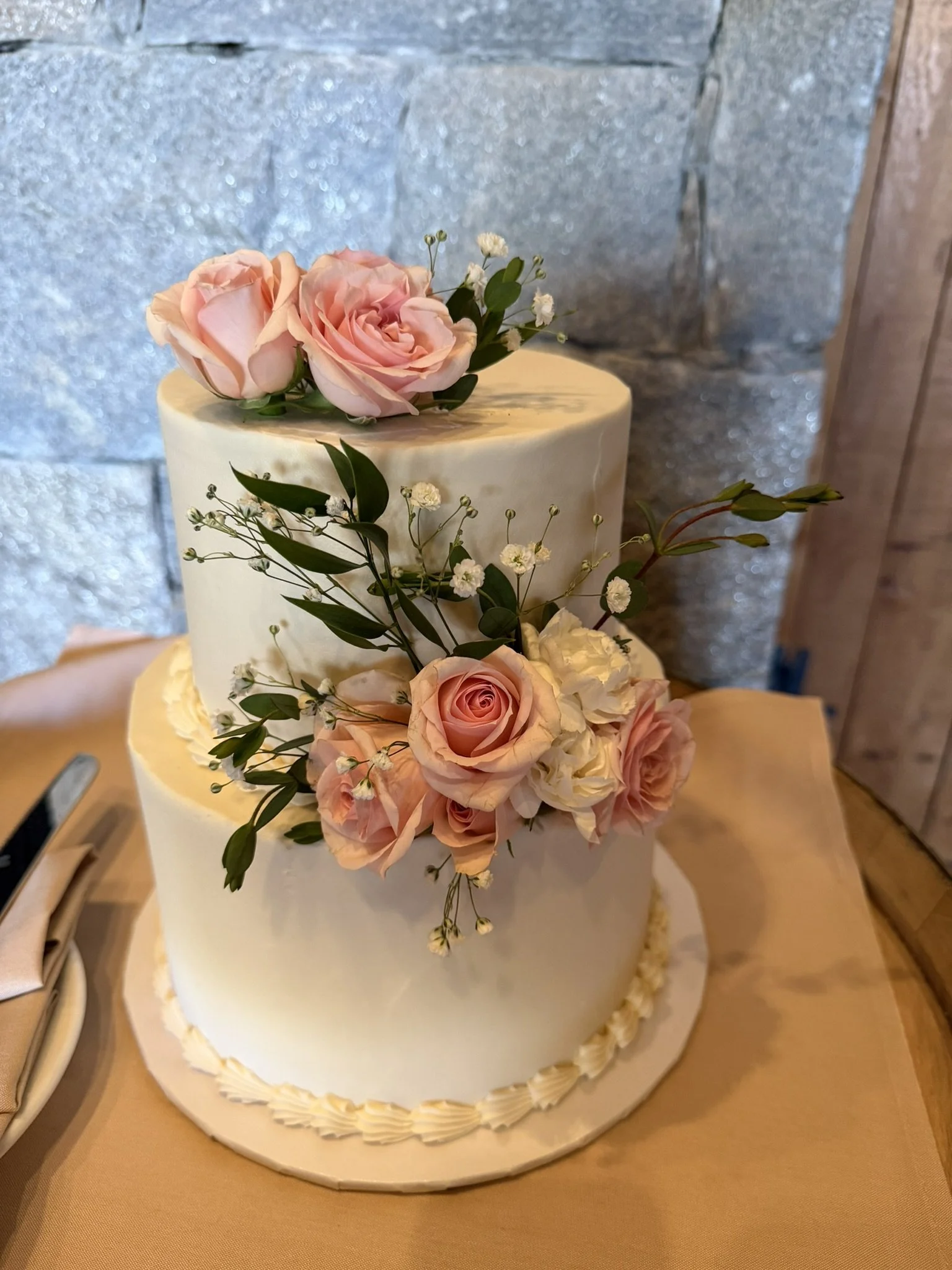 A two-tier white wedding cake decorated with pink roses and small white flowers with green leaves.