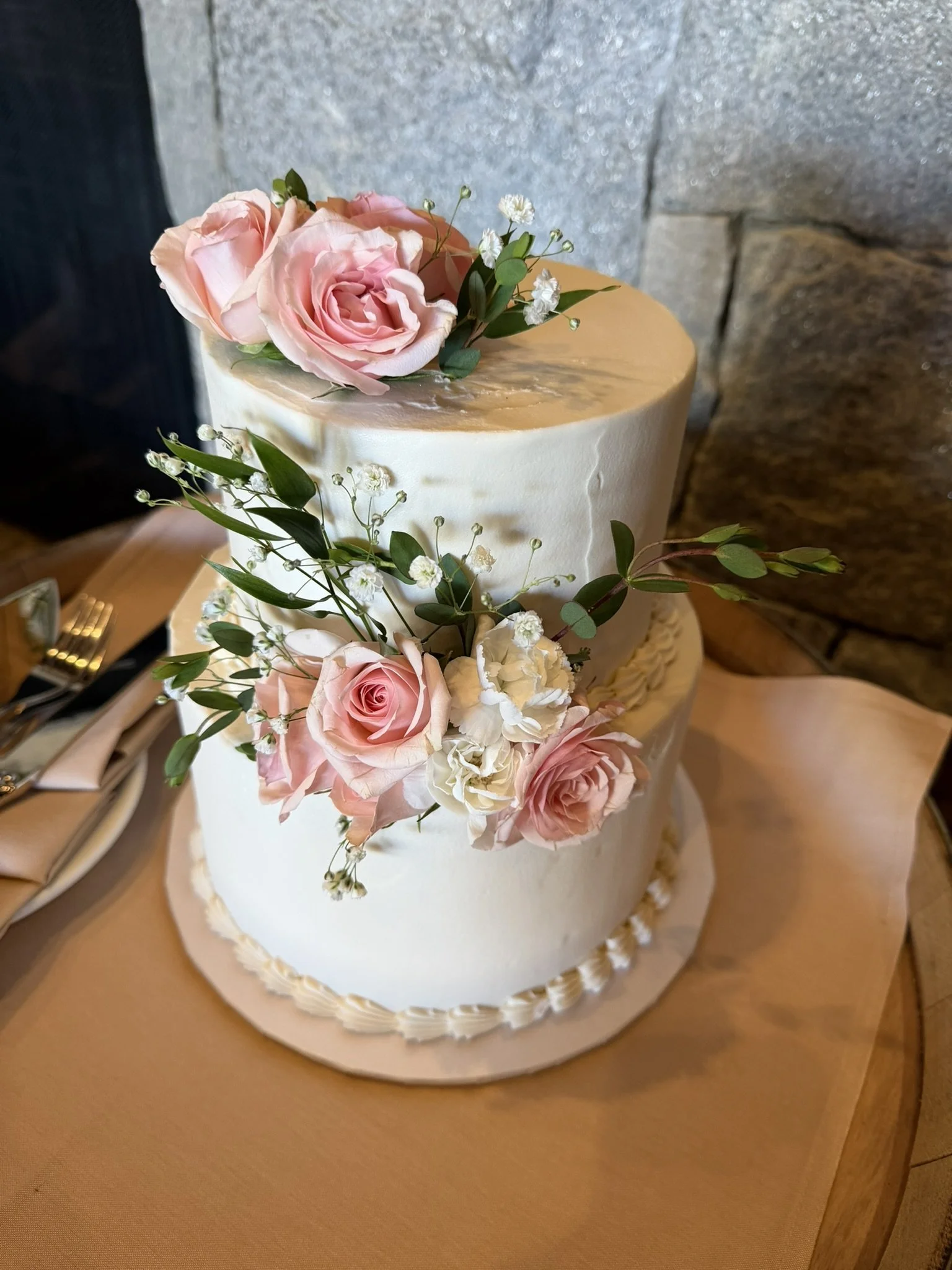 A tiered wedding cake decorated with pink roses, white flowers, and greenery on a wooden table.
