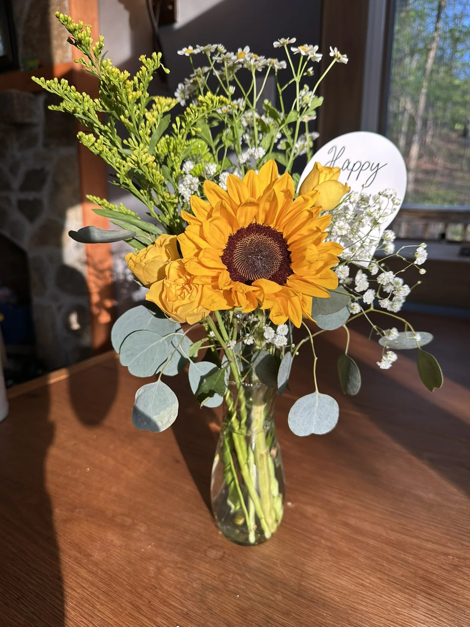 A clear glass vase containing a bouquet of bright yellow sunflower, white baby's breath, and green foliage on a wooden table.