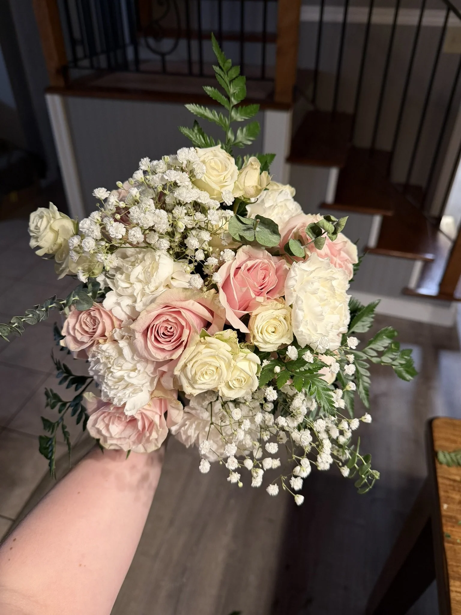 A hand holding a bouquet of flowers including pink roses, white roses, baby's breath, and green foliage, with a staircase and wooden furniture in the background.