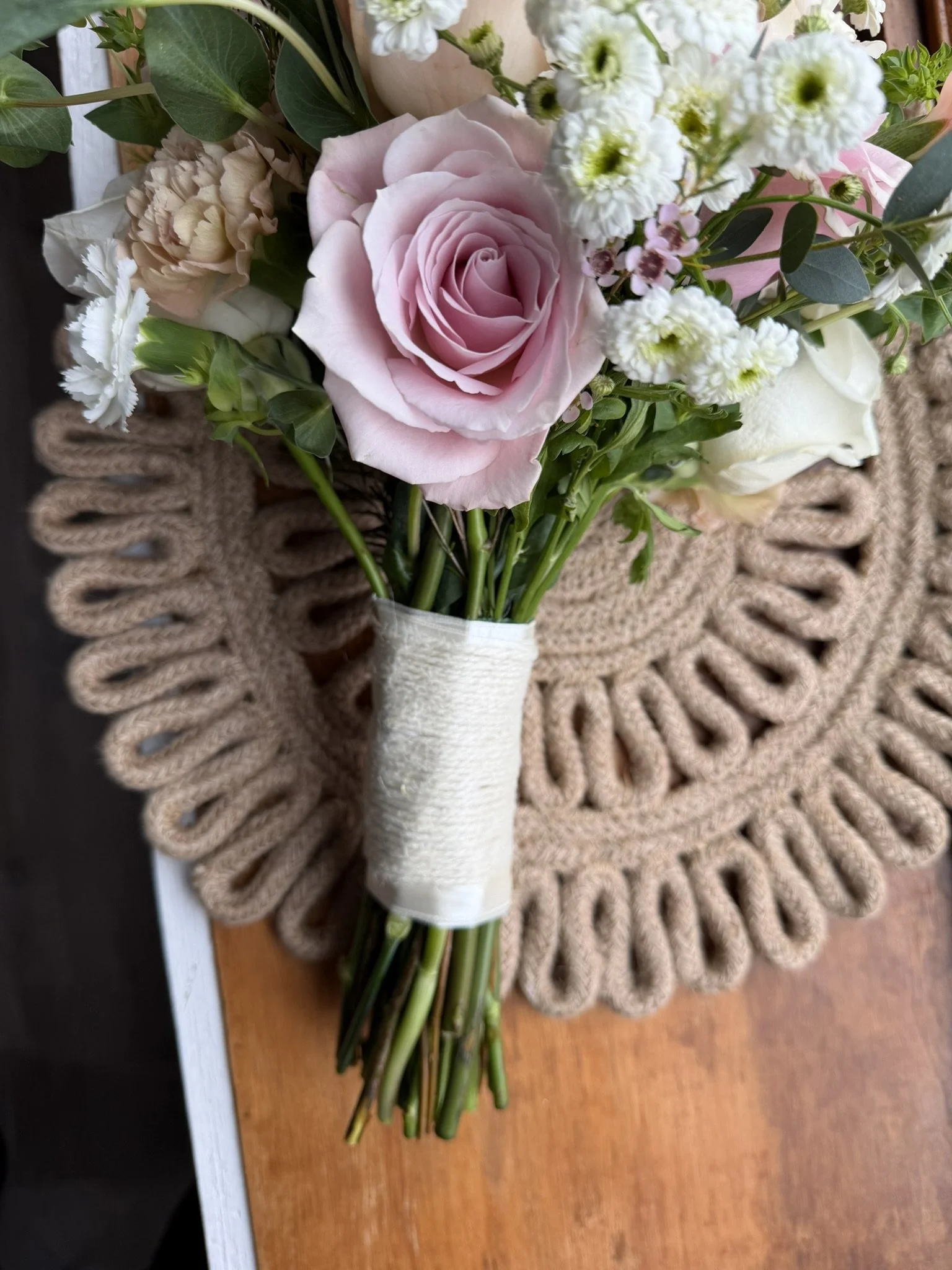 A bouquet of pink and white roses, white carnations, and other small white flowers with green leaves, placed on a circular woven beige placemat on a wooden surface.